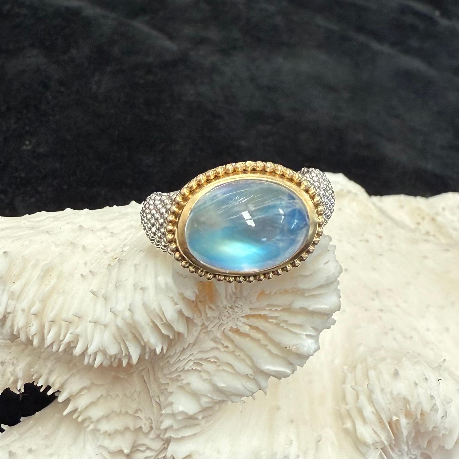 Ein sehr hochwertiger, blinkender 10 x 14 mm großer Regenbogen-Mondstein-Cabochon ruht in einer dicken, horizontal geschwungenen Fassung aus 18-karätigem Gold mit einem großen, umlaufenden 