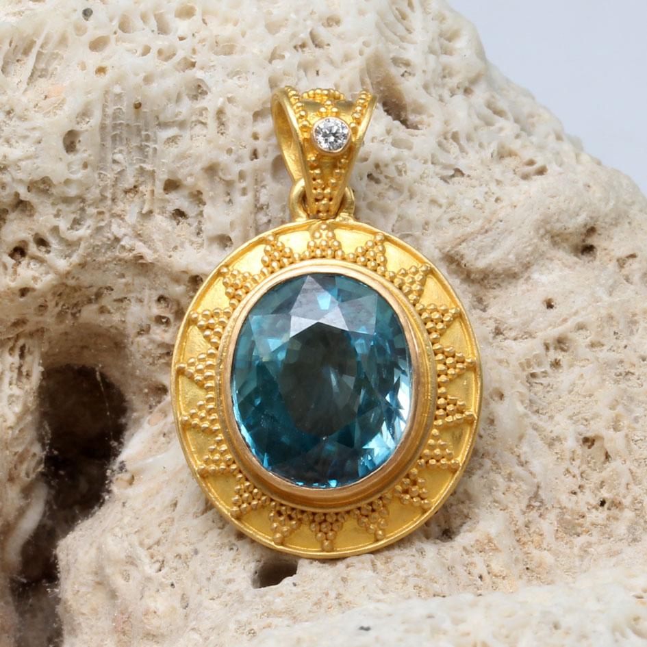 A highly lustrous 10 x 12 mm oval faceted Cambodian blue zircon is centered in an artisanally handcrafted high-karat gold oval setting with stacked 