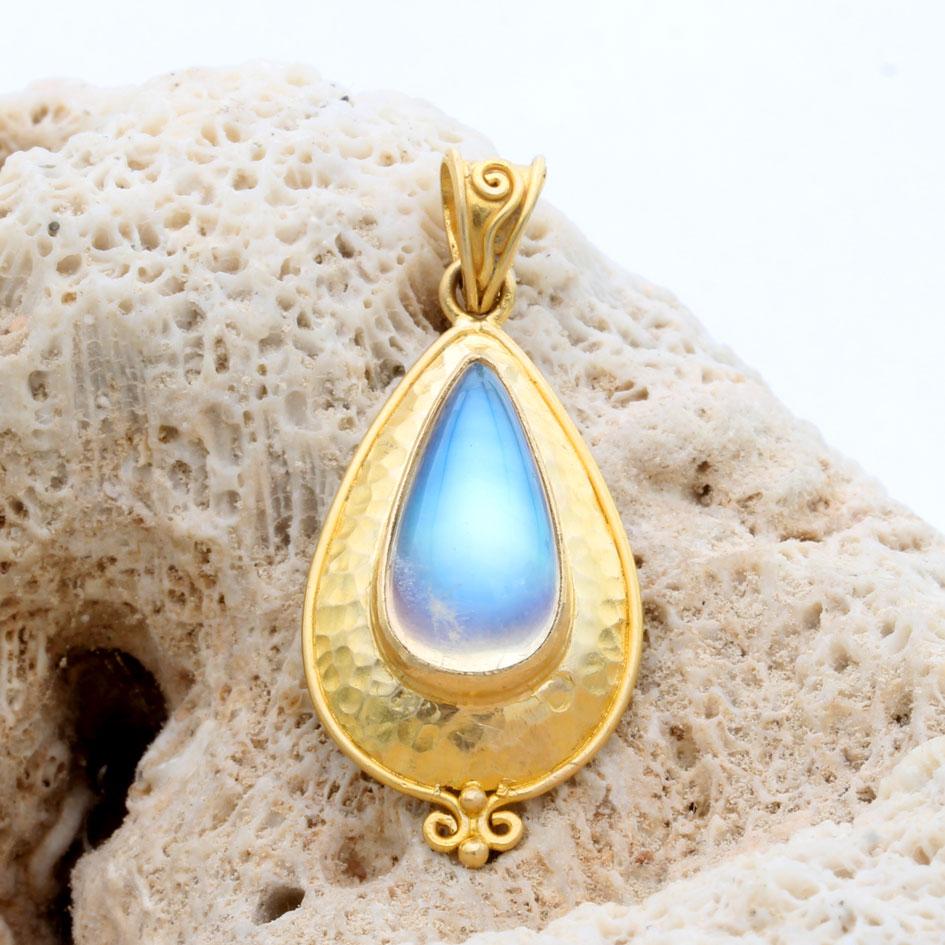 Un cabochon de pierre de lune arc-en-ciel en forme de poire de 8 x 17 mm de long repose dans une gloire d'or 18K martelée avec une délicate bordure de fil métallique se terminant par une granulation 