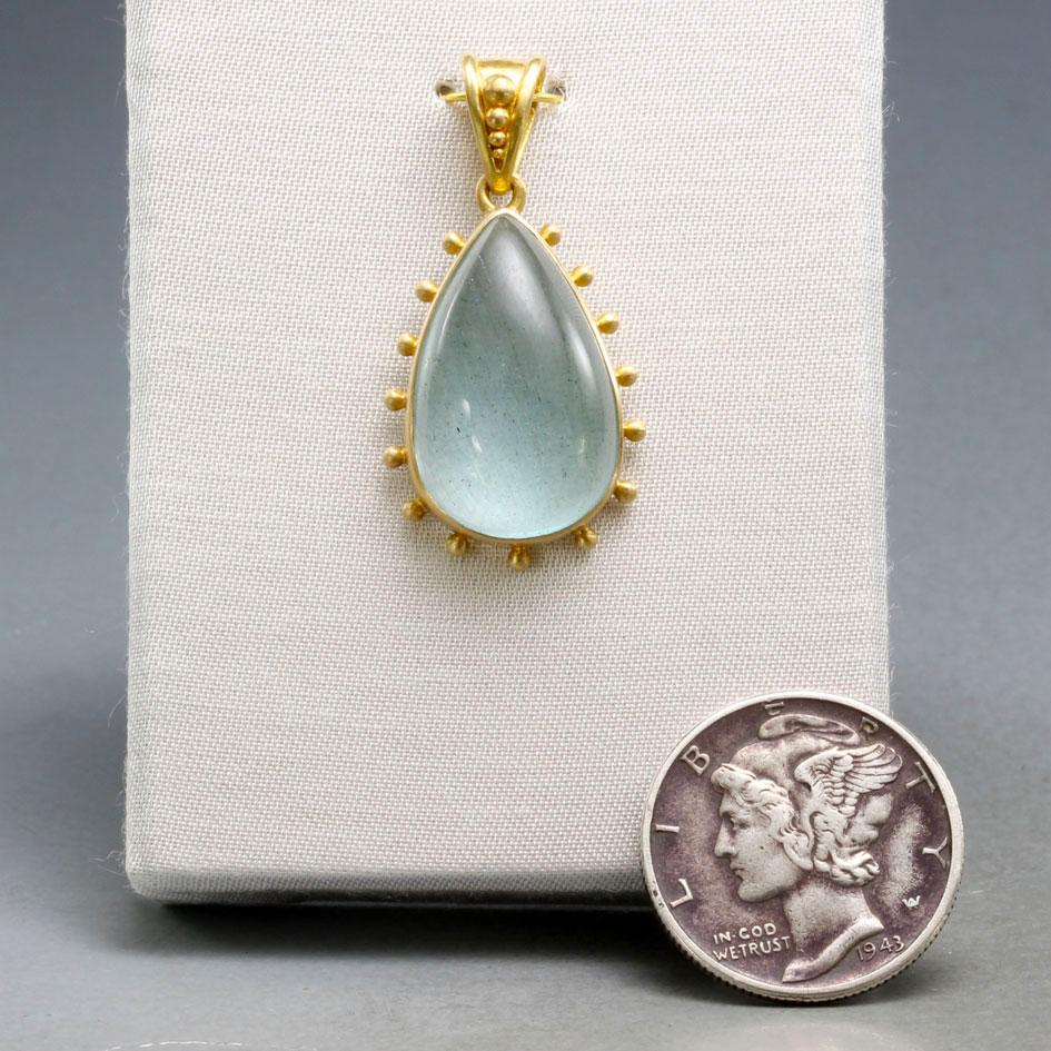 A limpid 12 x 18 mm pear shaped aquamarine cabochon is set in 18K matte-finish gold with evenly spaced large 