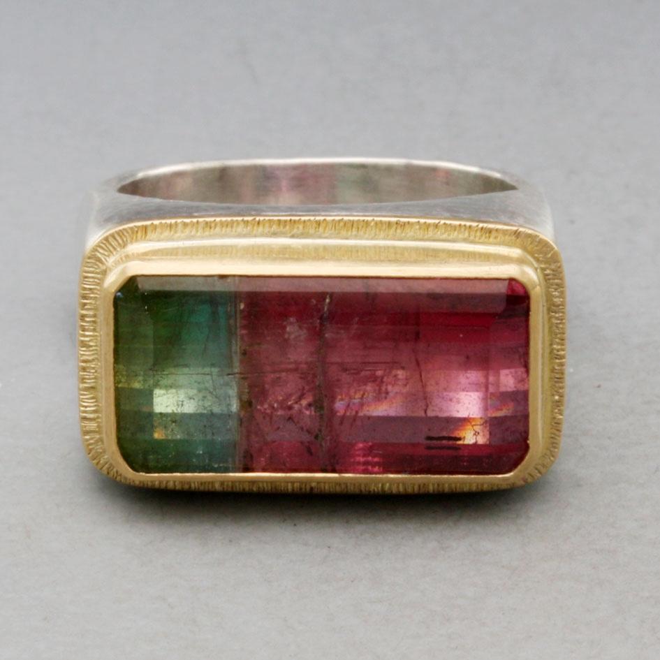 A bright and lively Brazilian 10 x13 mm octagonal faceted tourmaline displaying beautiful gradiation between light greens and pinks is set horizontally surrounded by a line texture accented 18K gold bezel, all atop a substantial 