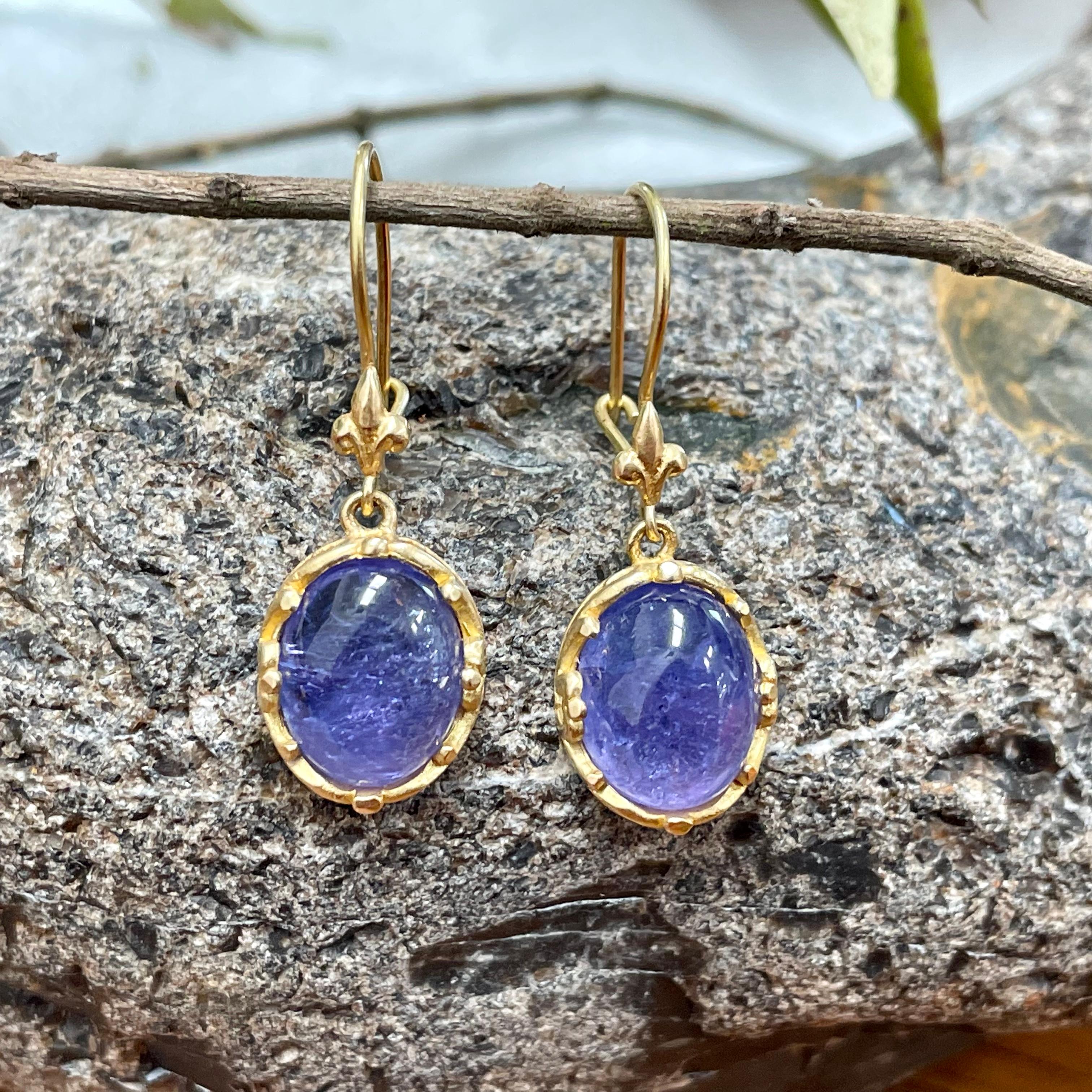Two 8 x 10 mm oval tanzanite cabochons are held in delicately carved bezels below fleur de leu ornamented 18K safety clasp wires. Beautiful contrast between the purple/blue of the stones and the gold. 