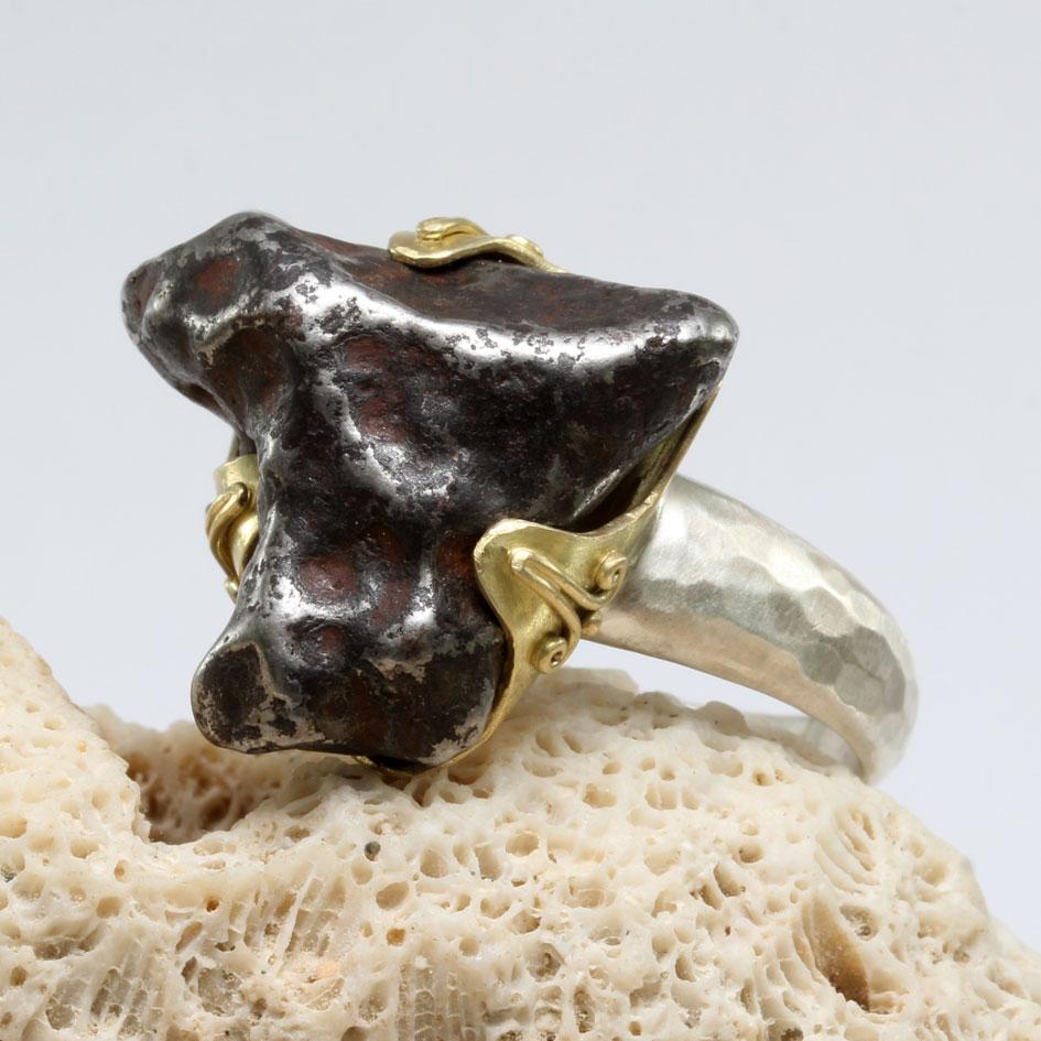 Ein großes, dreieckiges Fragment des Alin-Meteoriten wird in diesem Herrenring-Design von einer 18-karätigen Goldumarmung auf einem spitz zulaufenden und gehämmerten Sterlingsilber-Schaft gehalten.  Die 3 Zacken der Goldfassung sind leicht mit