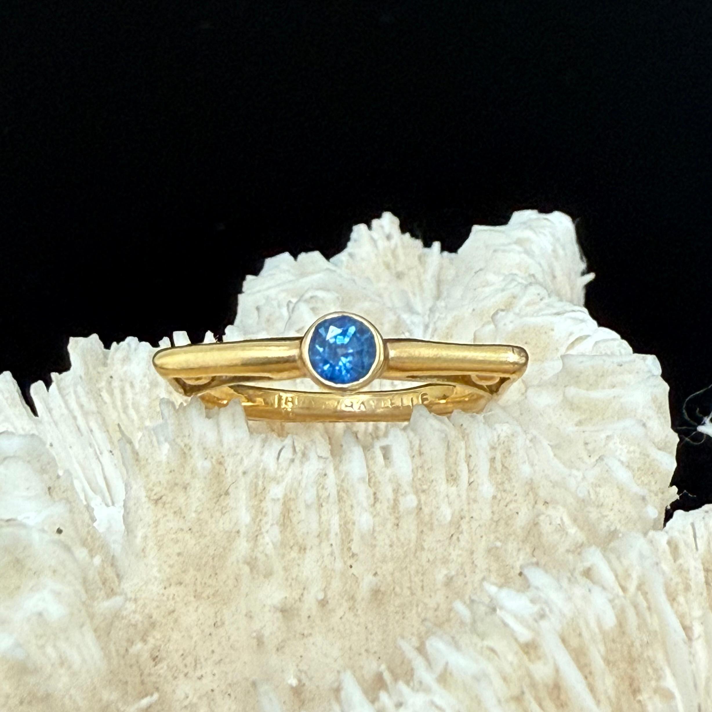 A bright 4mm round faceted blue sapphire rests centered within a unique wide topped matte-finish 18K gold band shaped somewhat squarely with four 