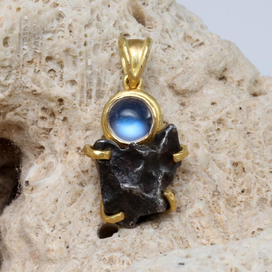 Ein unregelmäßiges 7 x 11 mm großes Fragment eines Sikhote-Alin-Meteoriten wird von 18-karätigen Goldzacken unter einem 1,0-karätigen, 6 mm großen, blau schimmernden Regenbogen-Mondstein gehalten, der von einer mattierten, gehämmerten 18-karätigen