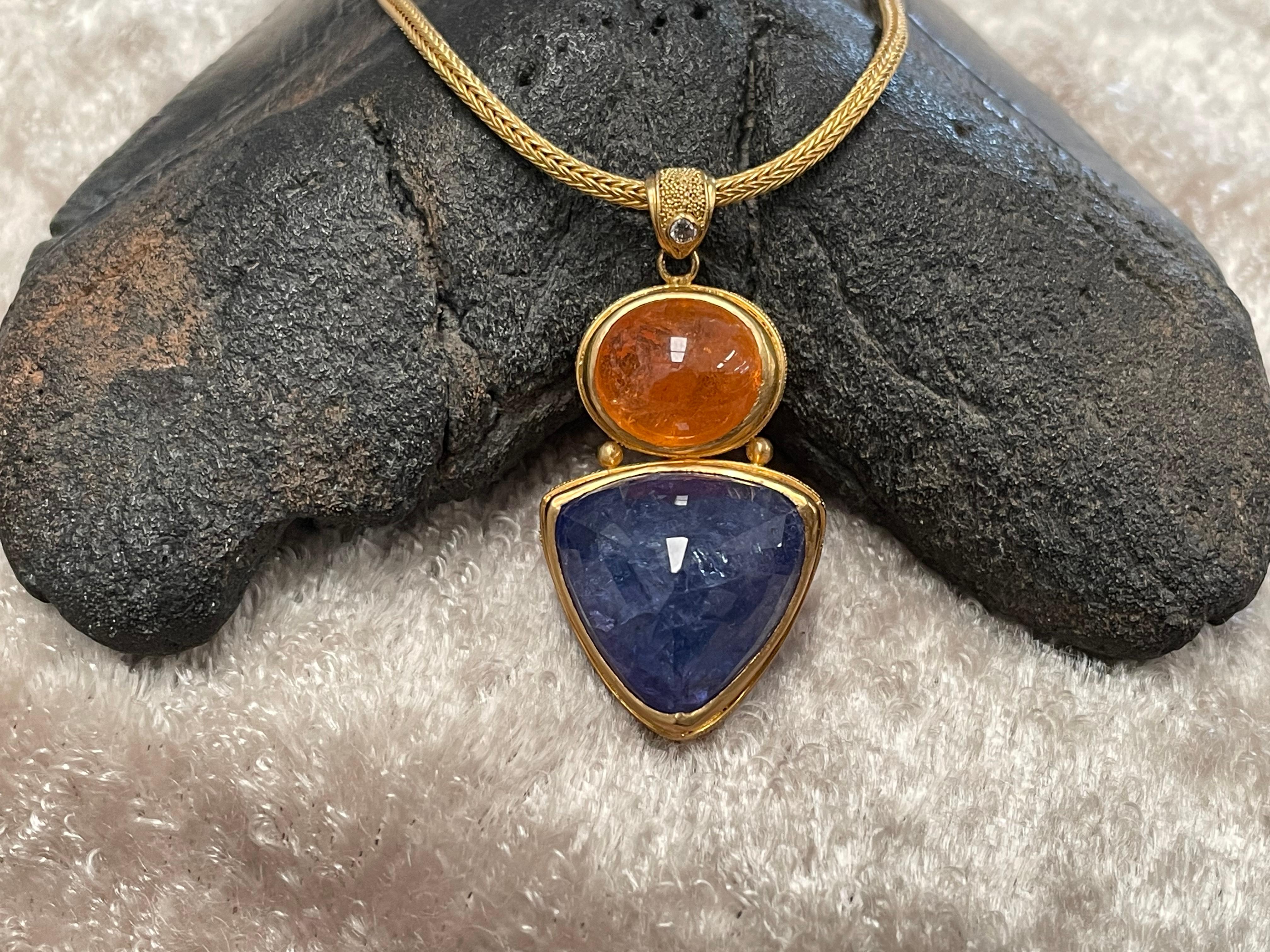 A 25.9 carat faceted 14 mm trillium Tanzanite is suspended below a horizontally set 11 carat 8x10 mm oval spessartite garnet cabochon combined with a diamond accented bail, in this interesting juxtaposition of complementary colored stones. The