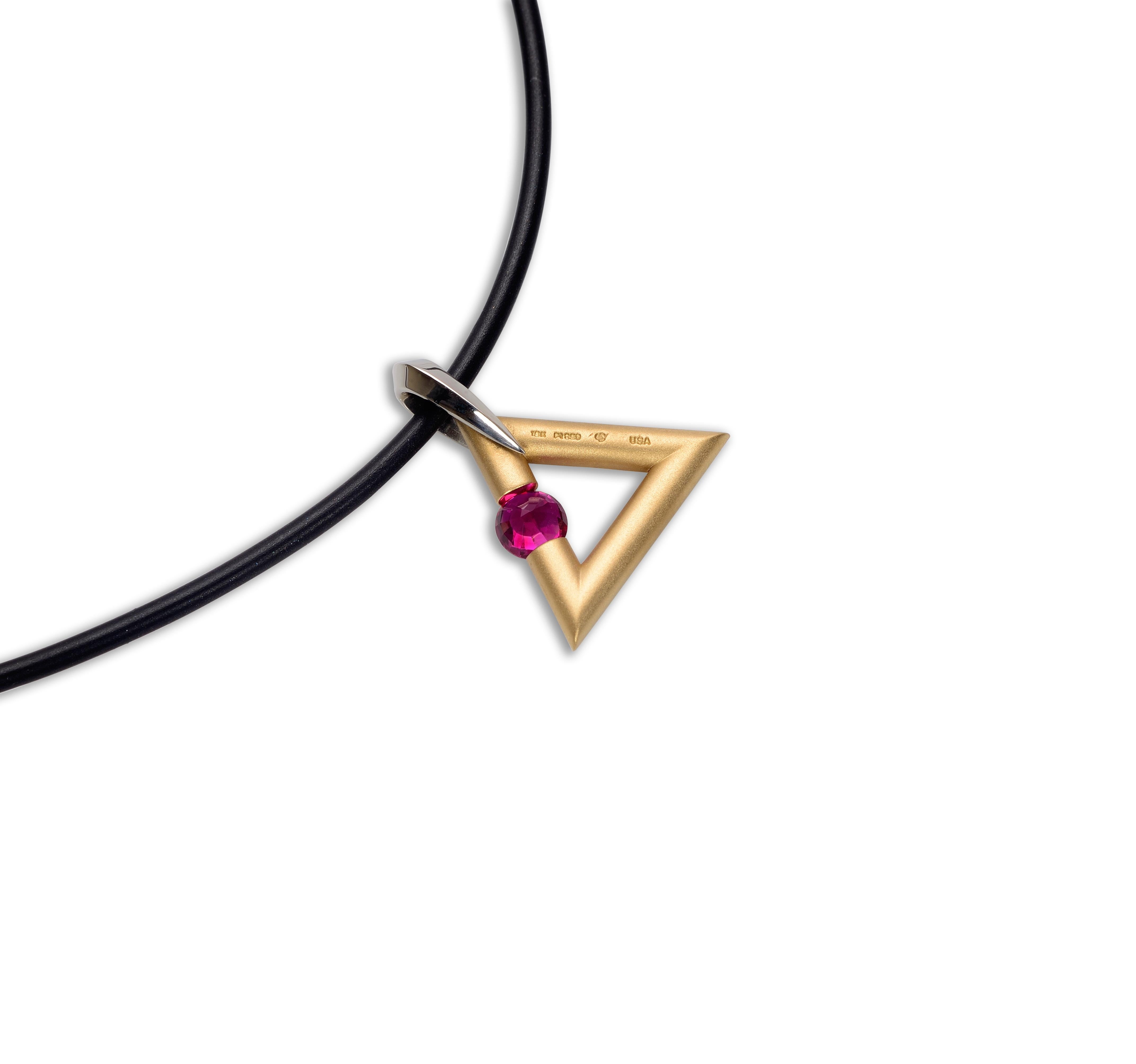 The Steven Kretchmer Logo/Triangle Shaped Pendant with a tension-set ruby is handcrafted in a matte 18K yellow gold with a shiny platinum bail. The reddish pink round cut 0.63ct ruby is tension-set in the side of the triangle shape. This pendant is
