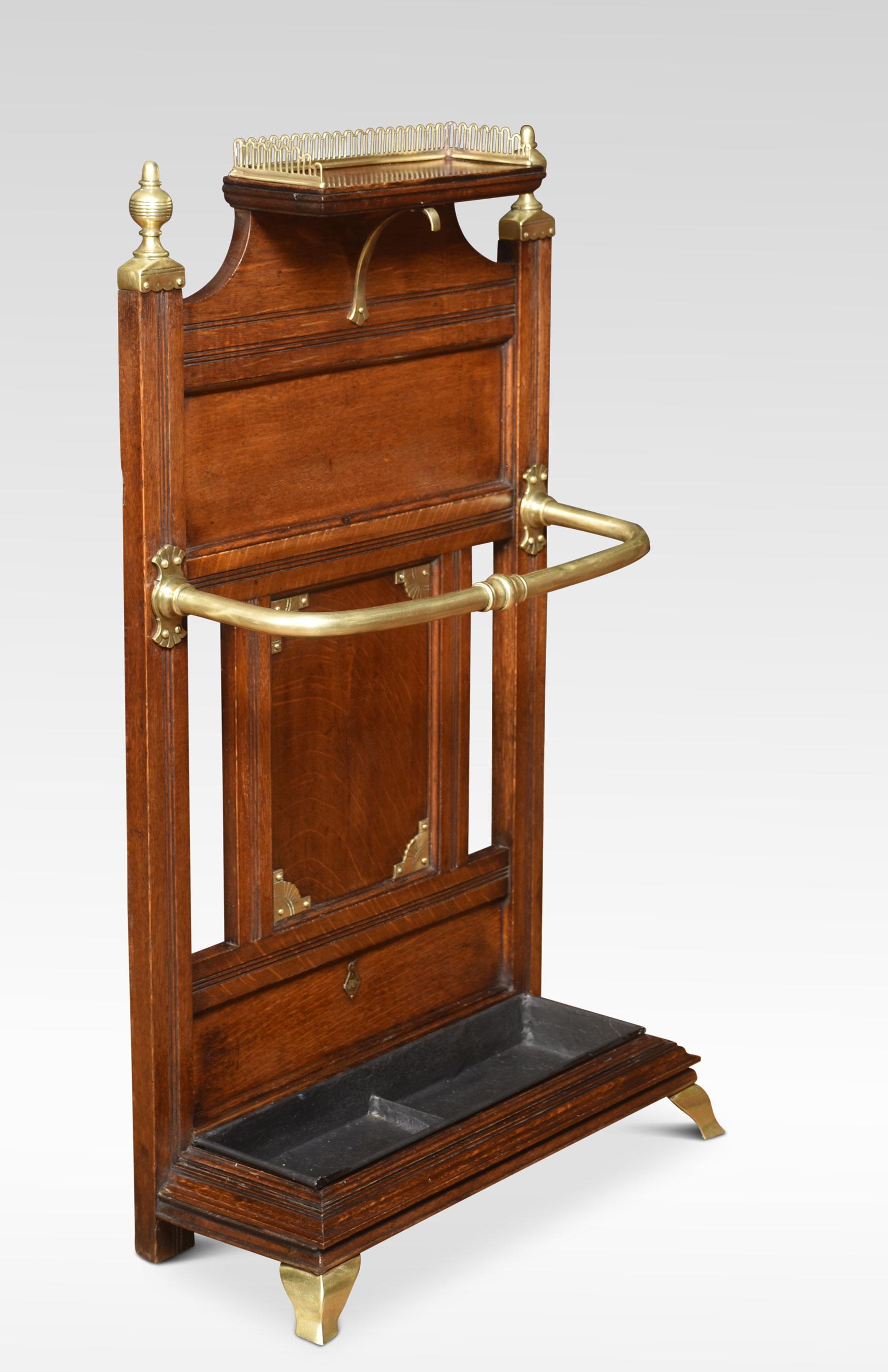 Oak and brass mounted stick stand by James Shoolbred and Co. Having raised three-quarter pierced gallery above a paneled back with turned brass finials and stick/umbrella holder to the molded base supporting a drip pan. All raised up on brass