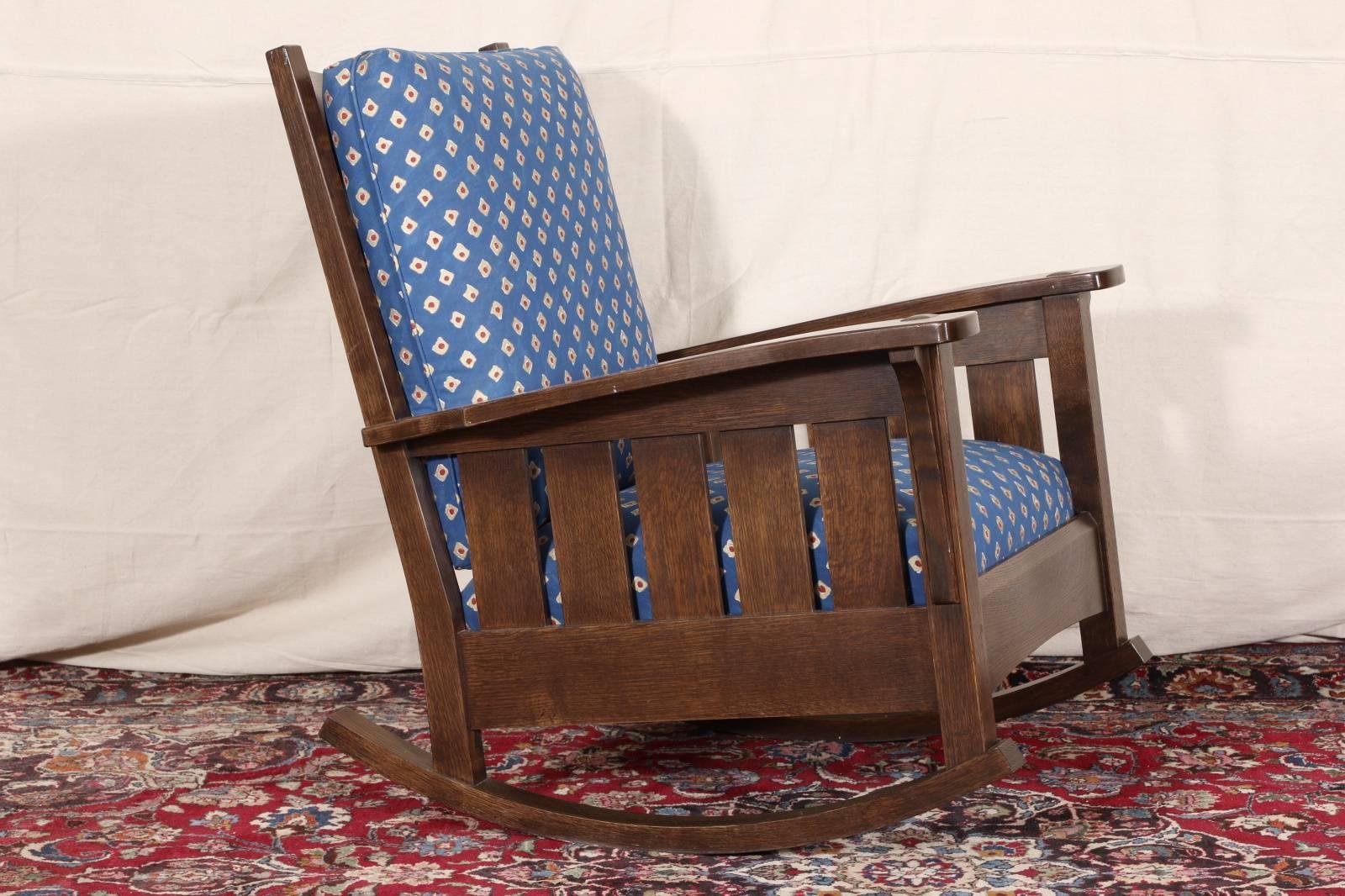 Stickley quarter sawn oak rocker, horizontal slatted back and vertical slatted sides, back and seat cushions upholstered in blue with white and red geometric pattern.

Condition: Expected wear and signs of use including some light surface