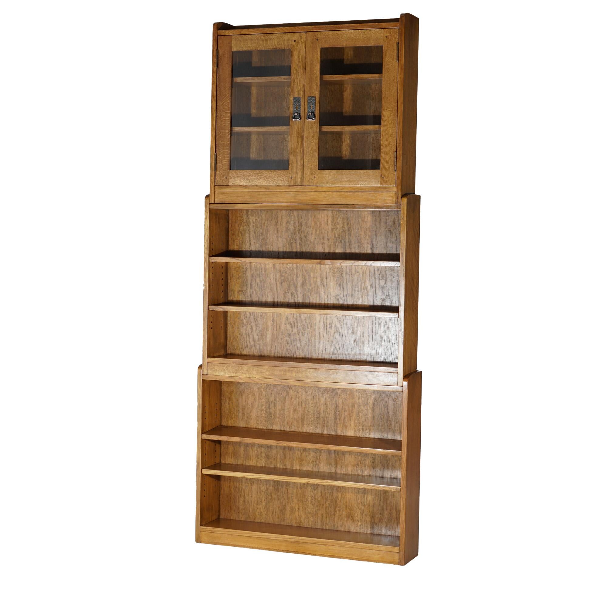 An Arts and Crafts style bookcase in the manner of Stickley offers oak construction in tiered modular form with three sections including two with open shelving and the top enclosed and having double glass doors; this item is designed to be used as a