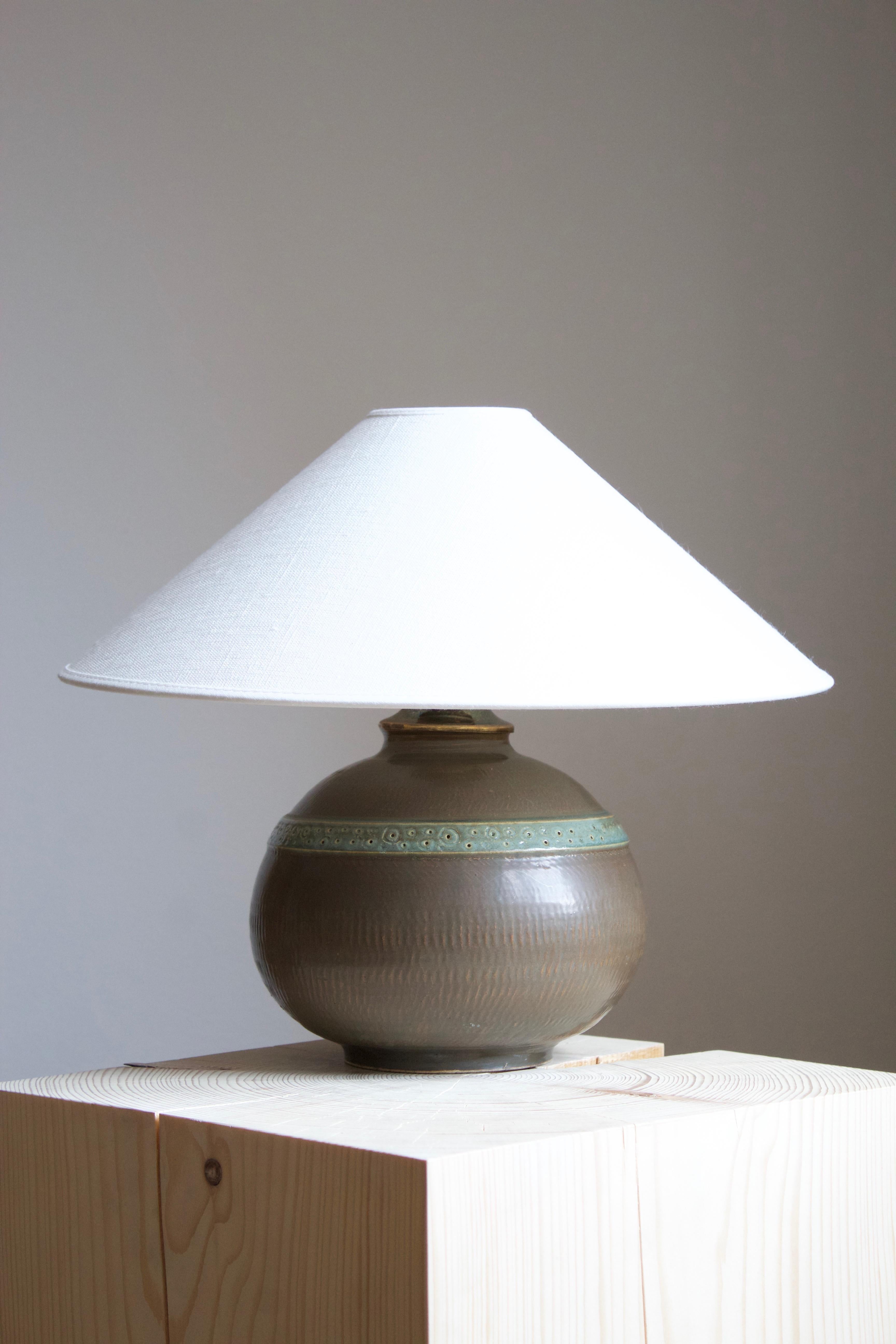 A table lamp or desk lamp designed and produced by Stig Lindberg. Produced by Gustavsberg. 

Marked with the Studio mark, separating this work from the large-scale production series by the maker.

Glaze features brown-green colors.

Stated