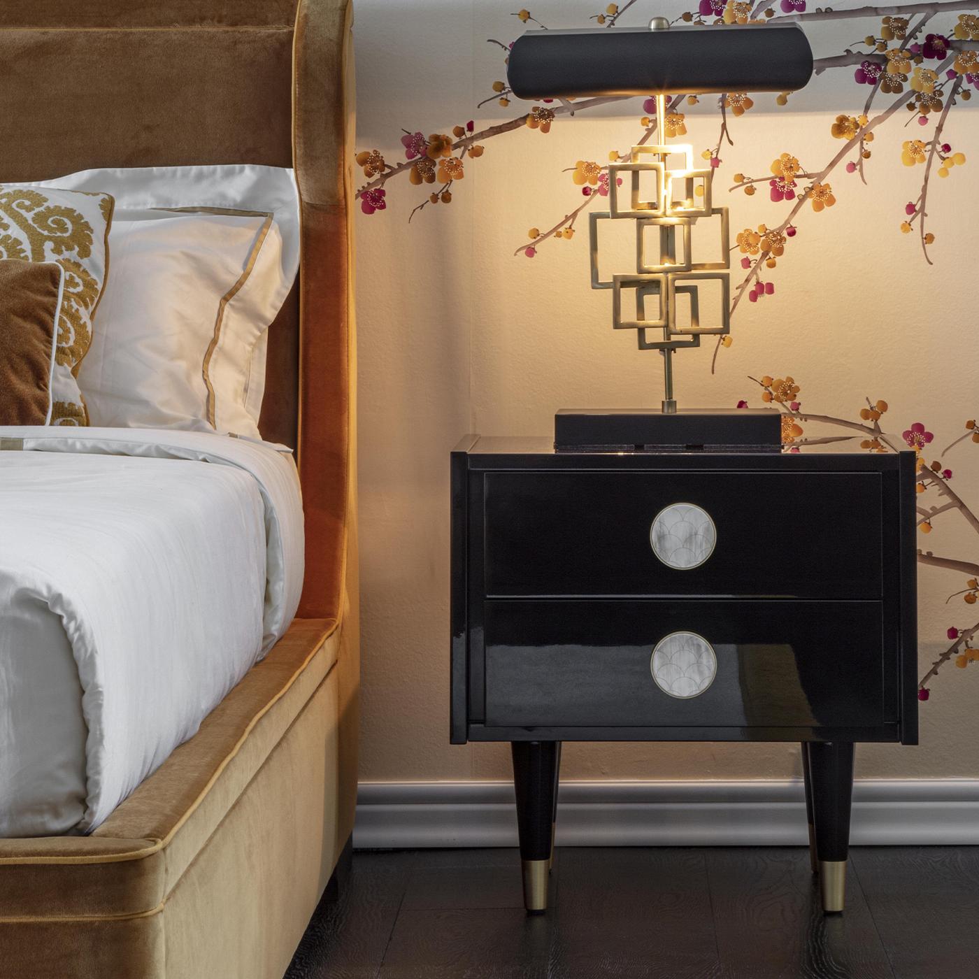 The sculpted wooden frame of this modern nightstand is lacquered in dark-brown with a glossy finish. The encased, two-drawer unit features front panels with round brass handles embellished with inlays in white buffalo horn. Paired with a twin to