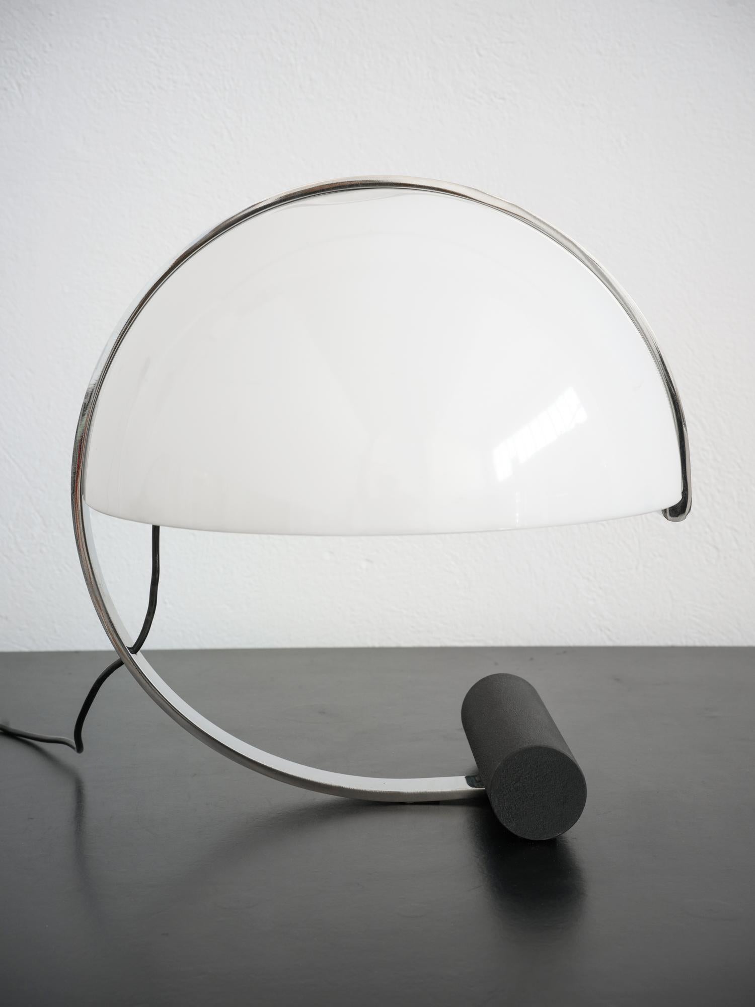 It is a Stilnovo table lamp characterized by a cylindrical base and an arched structure surrounding the diffuser, both made of metal. The diffuser in perspex, with a soft and rounded line, allows the light source to project a warm and intense beam