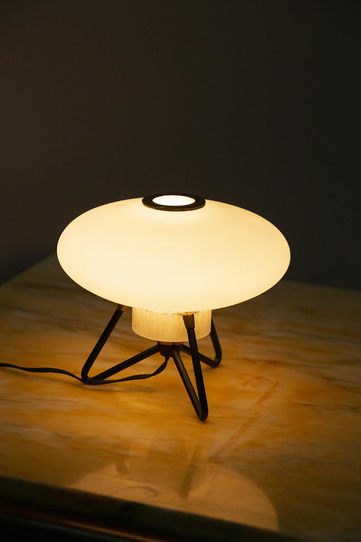 Stunning table lamp produced by Stilux in the 1950s. 
The lamp has a supporting frame made of black painted steel and brass in patina; the legs form a V so as to make it more durable.
Centrally arises the lamp cap in opaque opal glass. On the top