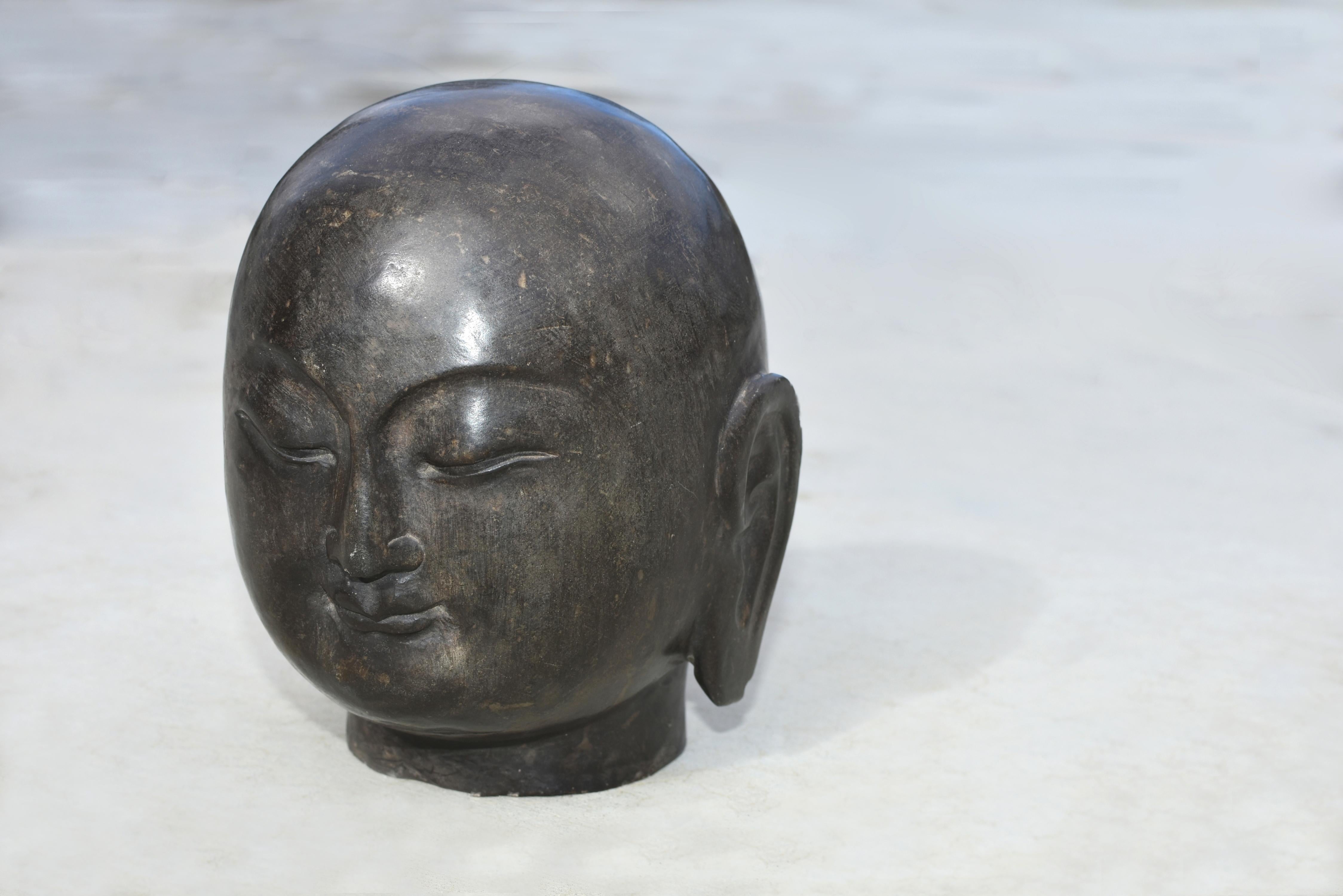 A beautiful, solid stone Buddha head. The round, full face is with downcast eyes beneath high arched brows above pursed lips, flanked by long pendulous ear lobes. The head hairless and perfectly smooth. The sculpture is Tang dynasty style, with the