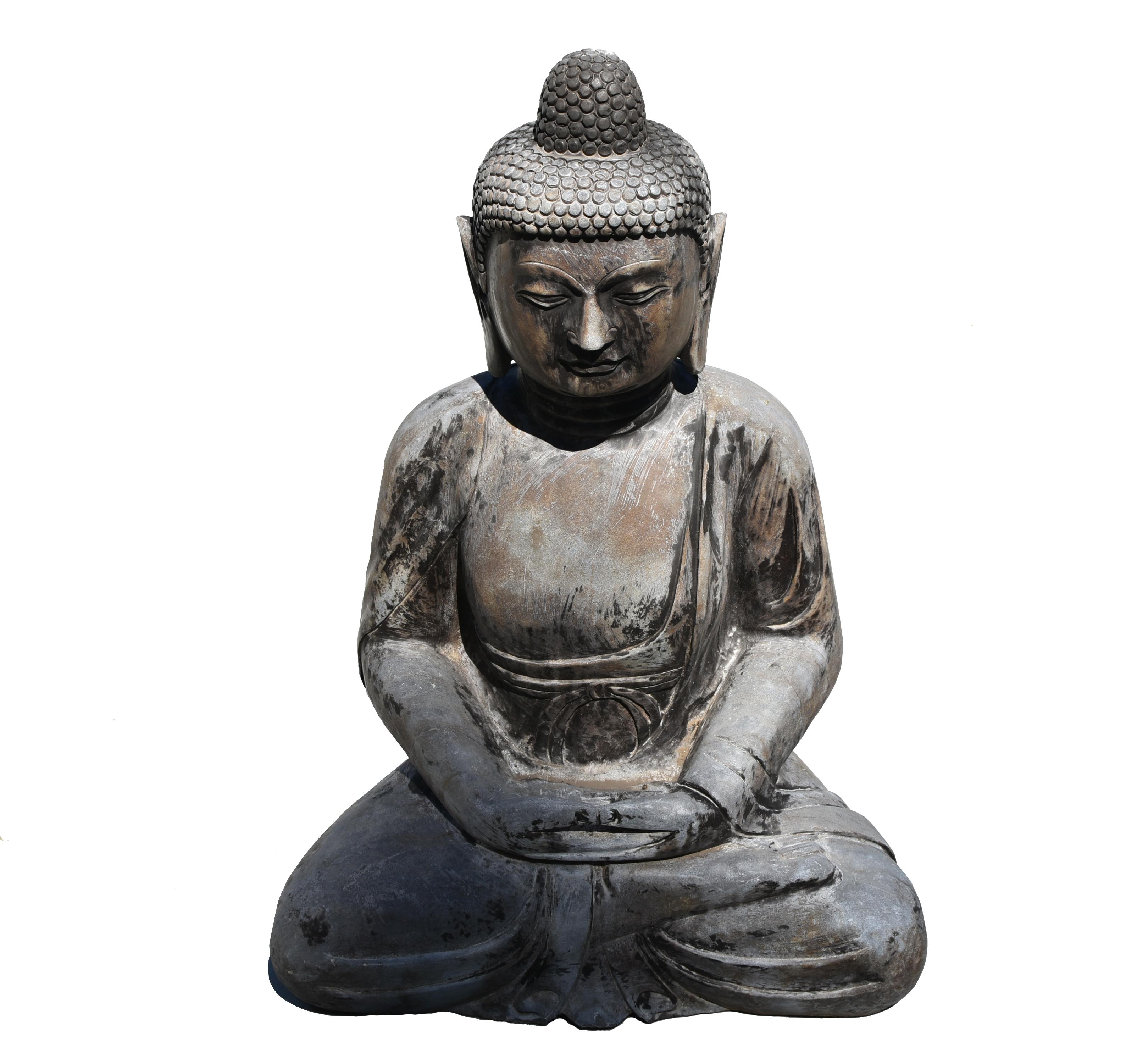 A large, solid stone, hand carved Buddha statue seated in lotus position. The broad face with high arched eyebrows and downcast eyes casting a serene aura, framed by long-lobed ears and beneath a pronounced hairline and surmounted by an ushnisha.