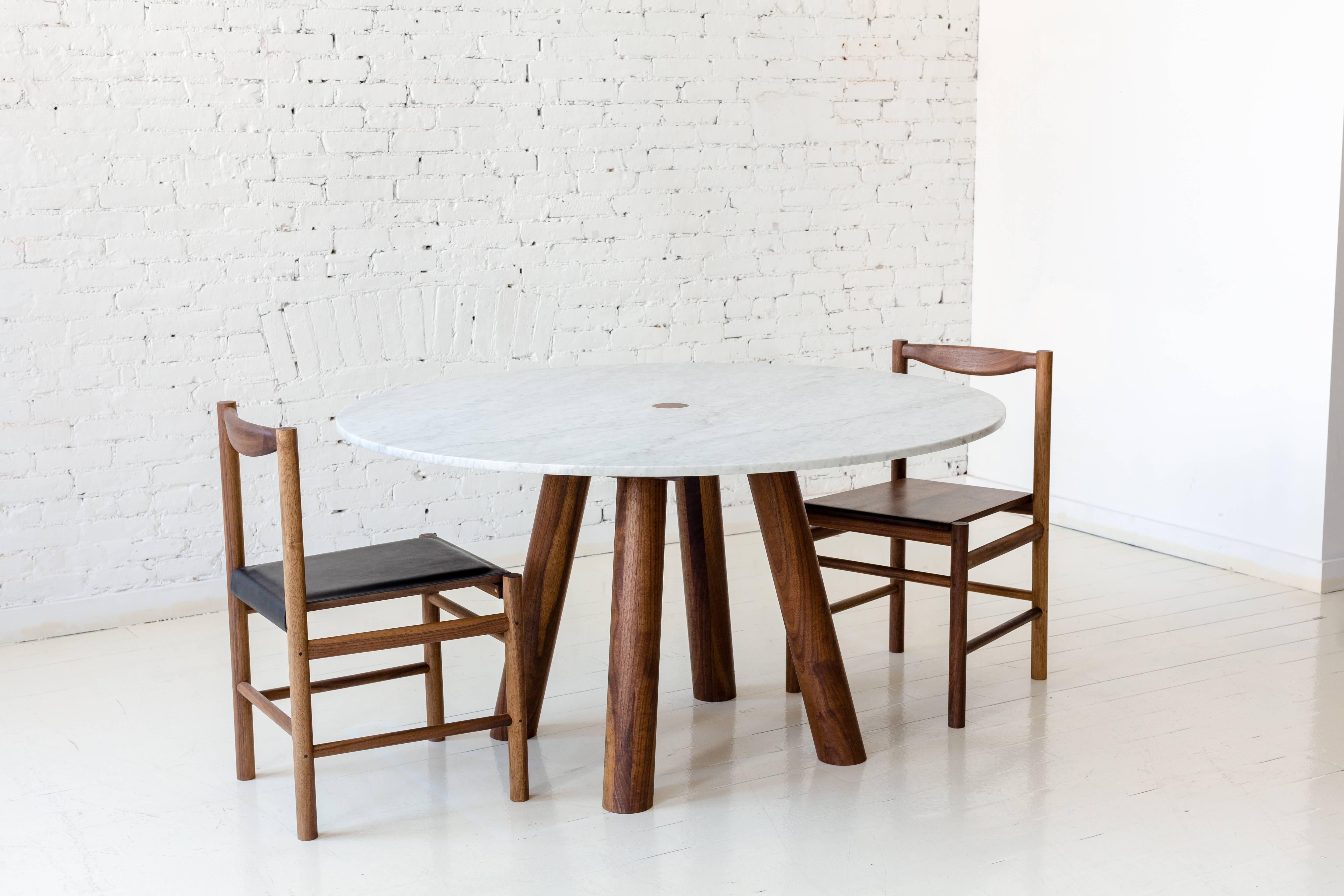 This contemporary, minimal dining table features large diameter round walnut legs and bold trestle supports to give rise to a white Carrara marble stone top. The table top features a wood surface detail that centers the top to the base and