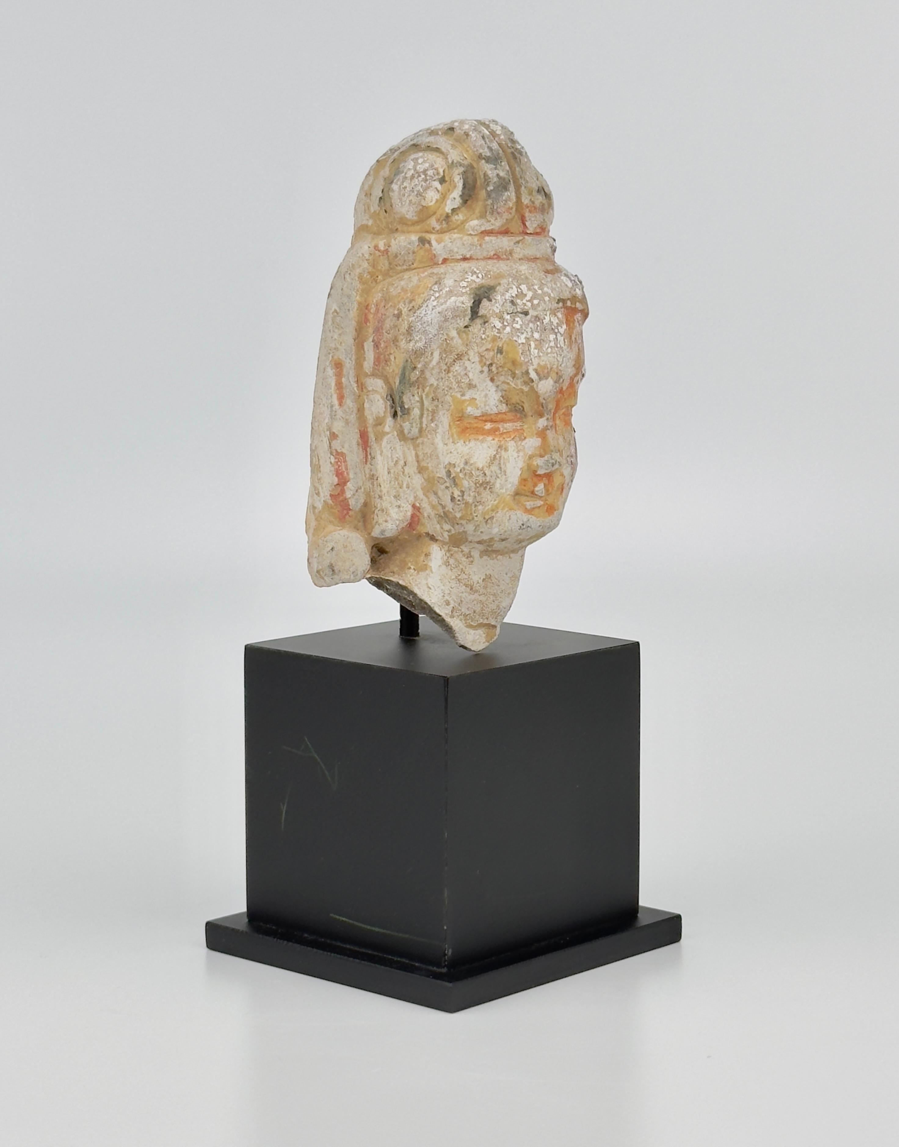 The face with downcast eyes and a smile, the hair swept up behind the pendulous ears into a high topknot. Faint traces of pigmentation hinting at the sculpture's original polychromy. The black stand was made in Hong Kong at the time of the