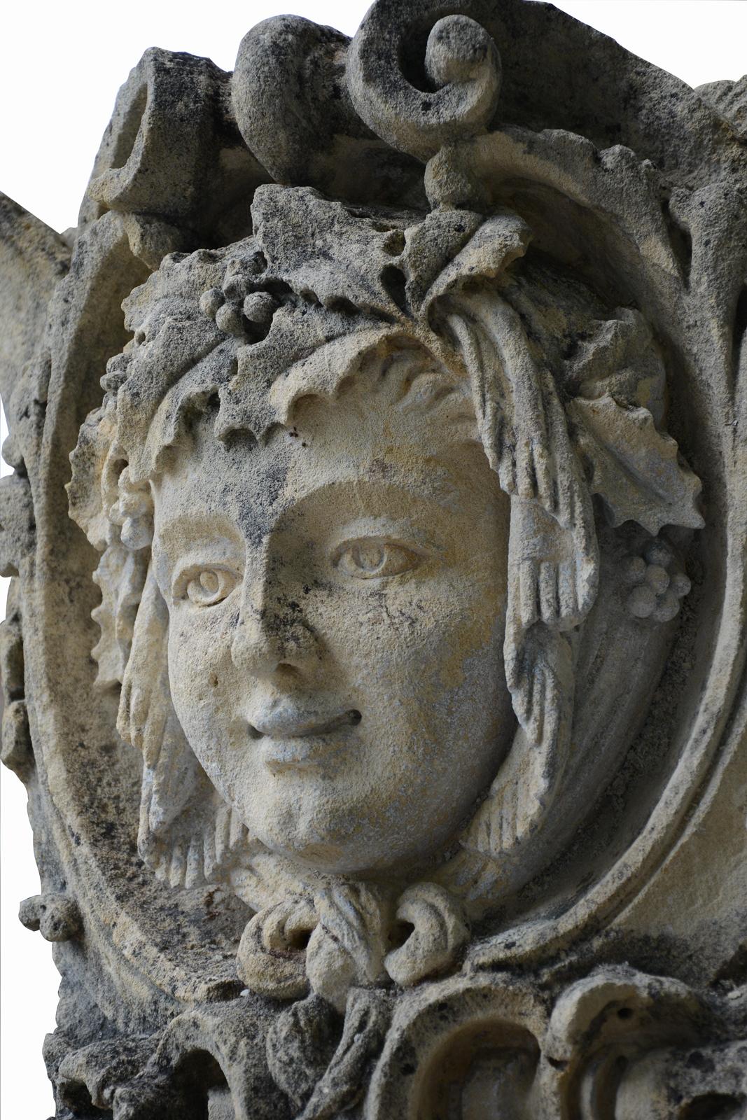 Dating from the 19th century, stone mascaron representing a woman. Its head is in a medallion formed of scrollwork and vegetable interlages. She wears a crown of holly and a scarf around her face and forming a knot in it. The holly is a plant very