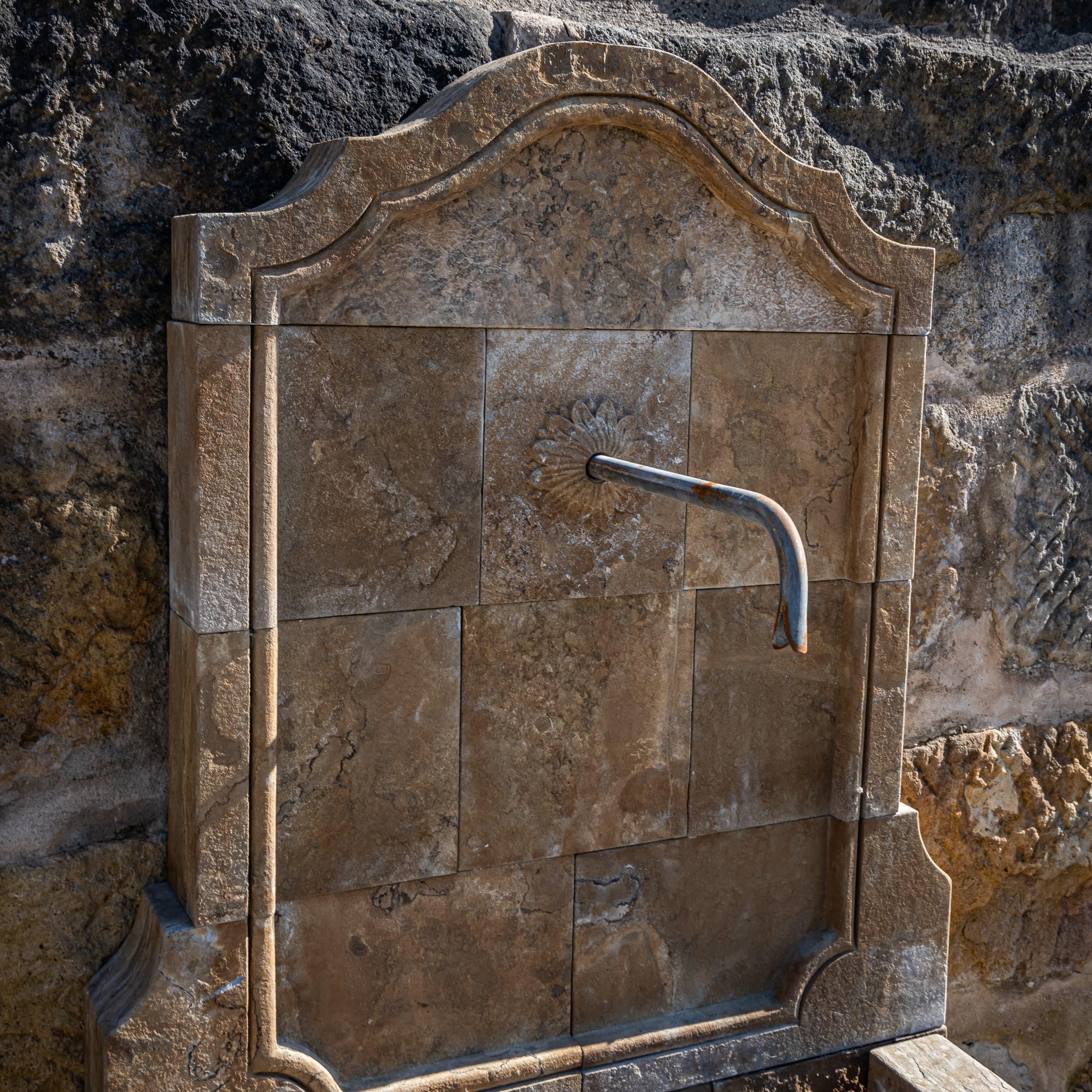 Wall fountain made of hand-hewn natural stone with a high, curved back panel and faceted basin. The harmonious arrangement of the stone slabs and the precisely crafted edges emphasize the artisanal quality and clean design language. 