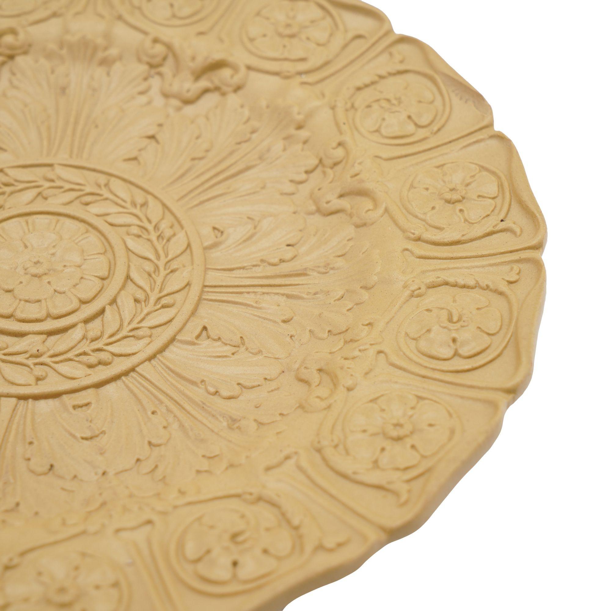 Putty colored stoneware plate with fine smear glaze. The impressed surface decoration centers on a Tudor rose with laurel leaf circlet, radiating acanthus leaves, and 16 compartmentalized scrolls, each featuring a single Tudor rose. The reverse is