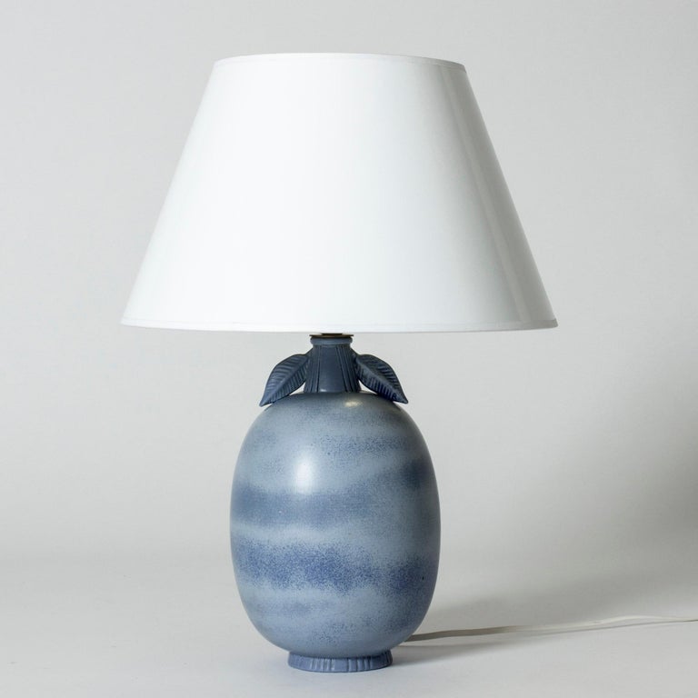 Lovely table lamp by Gunnar Nylund, made from stoneware. Plump, oval form with a decorative pair of leaves. Glaze blending pale and darker blues. Shade with golden inside that reflects a warm light.