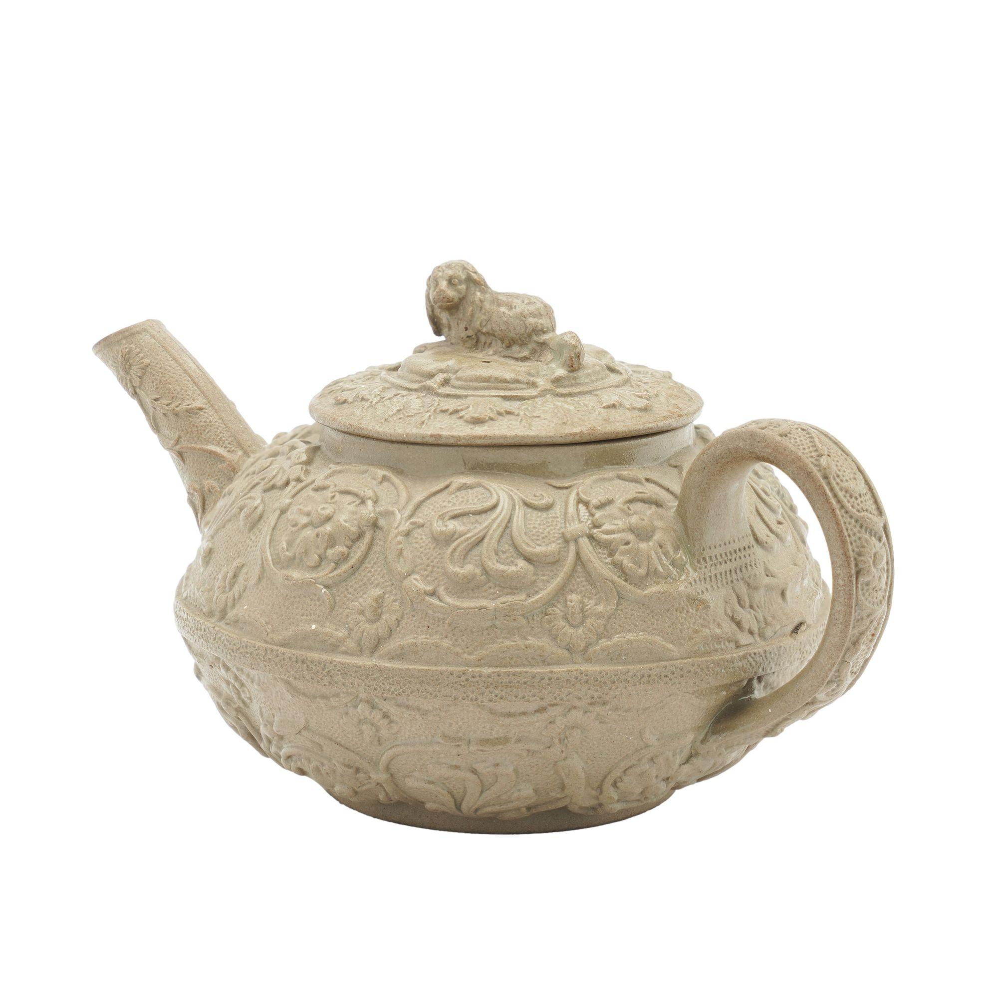 Slip cast stoneware tea pot in a putty colored smear glaze. The tea pot features a spaniel finial and heavy Renaissance scrolls of flowers on an overall dimpled ground. Cast from the Wedgwood & Bentley mold of 1775, which was discontinued after 1790
