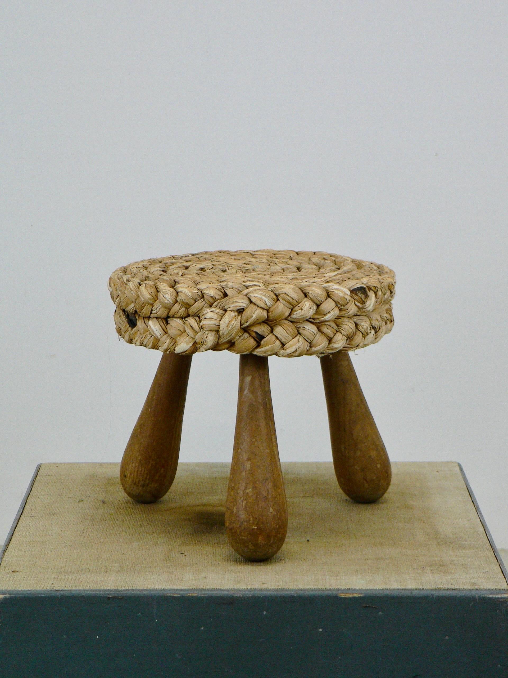 Mid-century French low Shepard's stool designed by Adrien Audoux and Frida Minet. This rustic French pouf showcases a circular seat crafted from woven raffia/rattan, supported by tripod legs and an oak keel base. Maintaining its original condition.