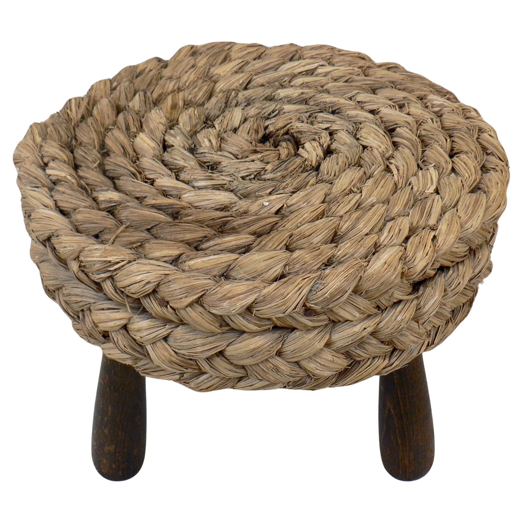 Mid-century French low Shepard's stool designed by Adrien Audoux and Frida Minet. This rustic French pouf showcases a circular seat crafted from woven raffia/rattan, supported by tripod legs and an oak keel base. Maintaining its original condition.