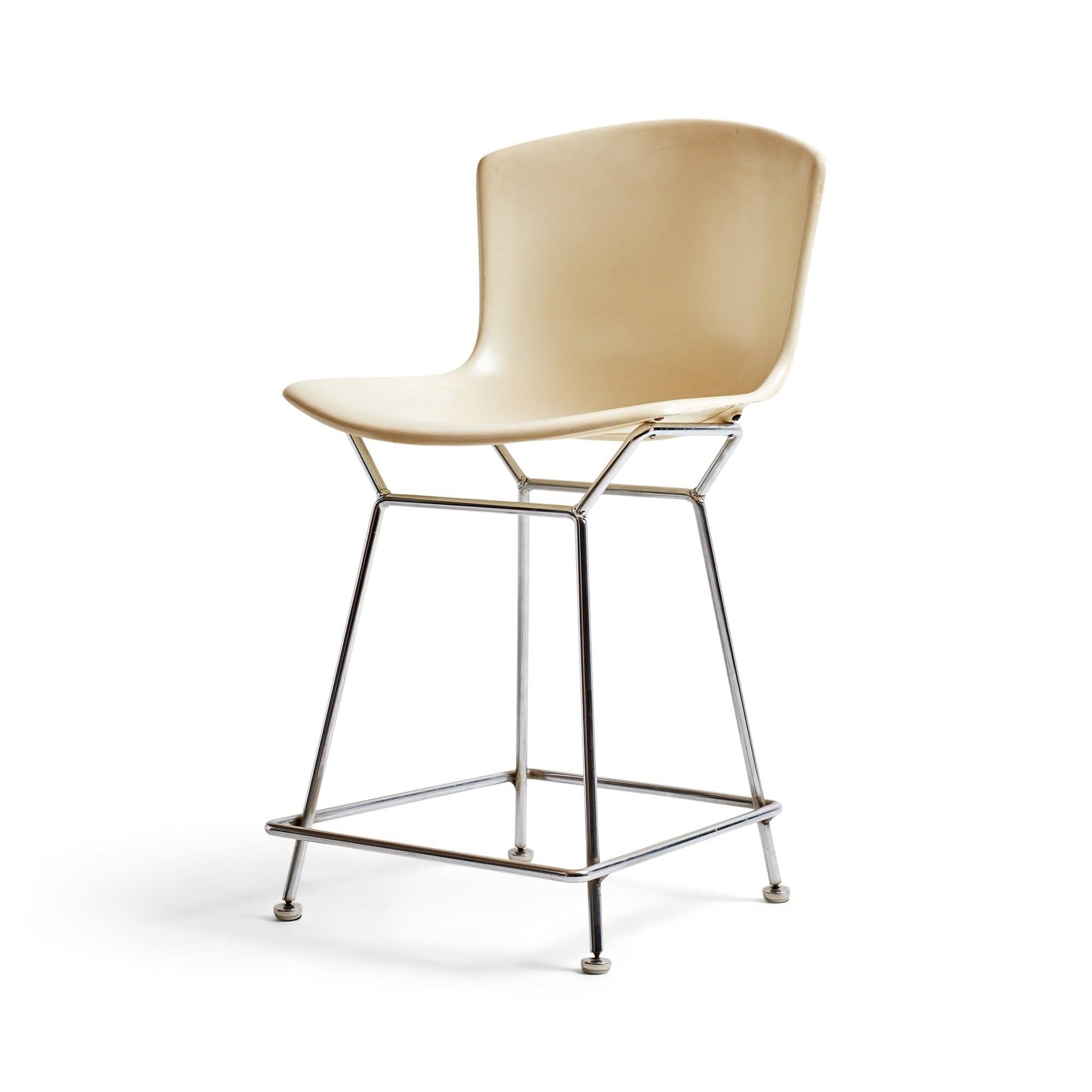 A counter-height barstool with a white plastic shell on splayed, chromed steel legs.