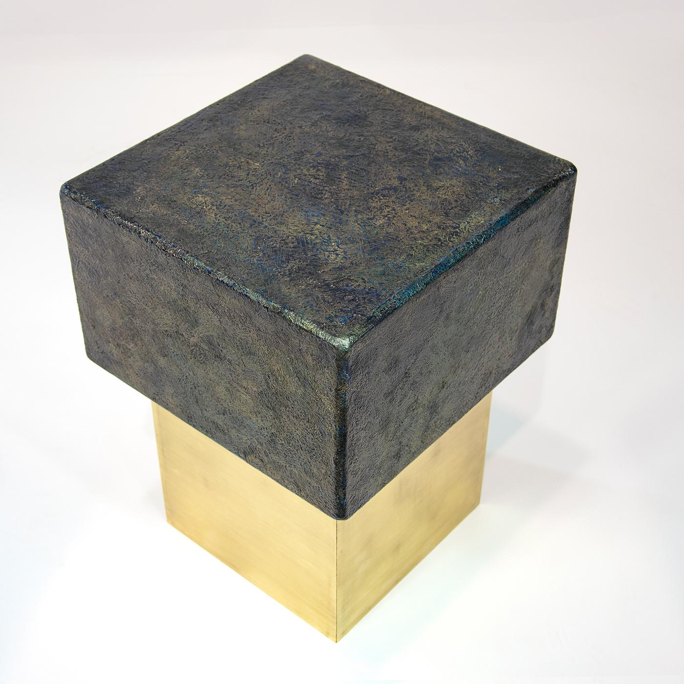 Wood and brass are the core elements of this stylish and contemporary small table, whose original design evokes the shape of a bottle stopper. Supported by a brass square base, the textured and metallic black patina finish of the square wooden top