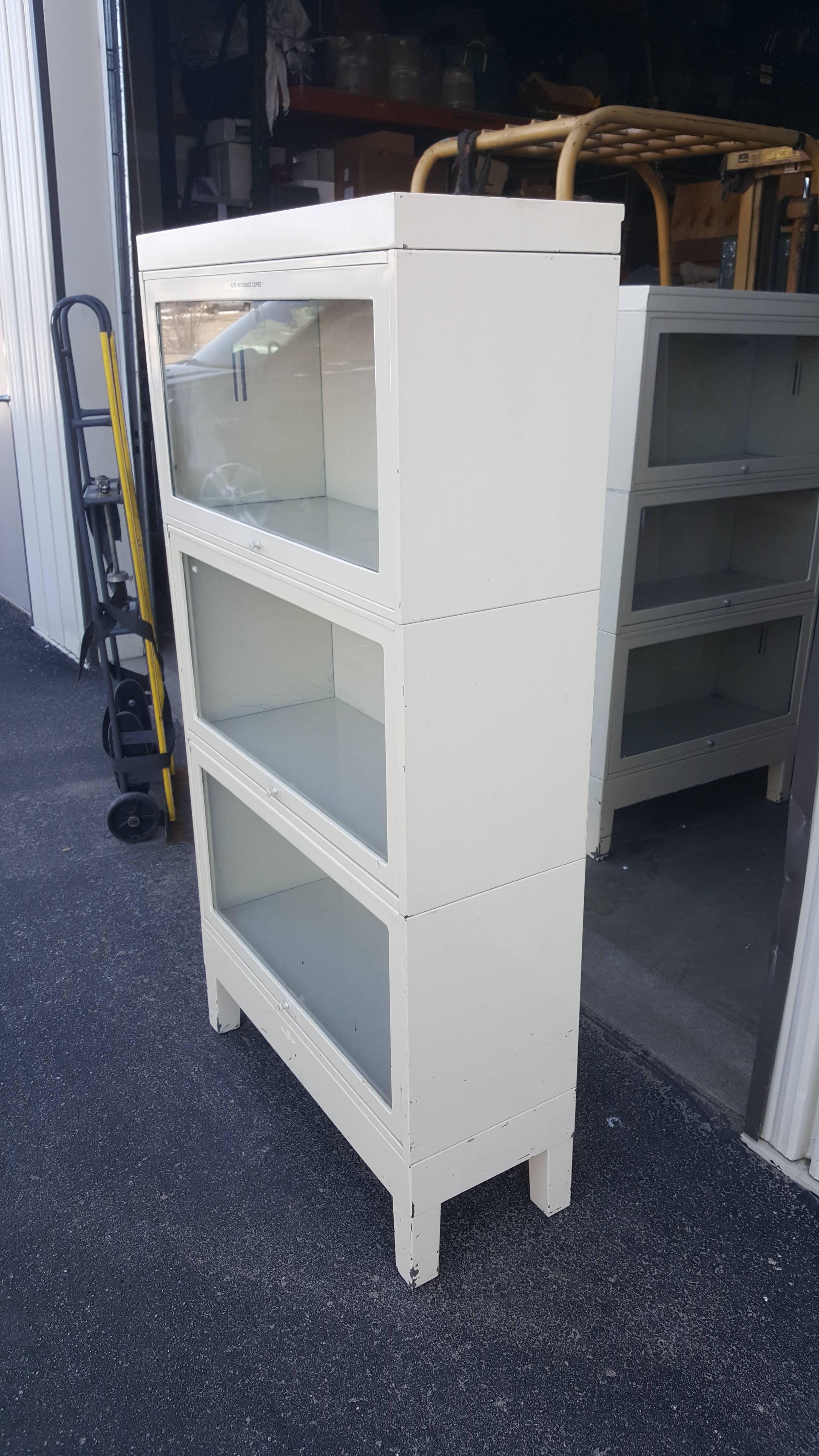 Barrister storage cabinet or bookshelf unit of painted ivory steel with glass fronts. Three stacking sections (and base), each with its own glass door front that slides up and under when open. Clean, sliding door fronts work perfectly. Usage: