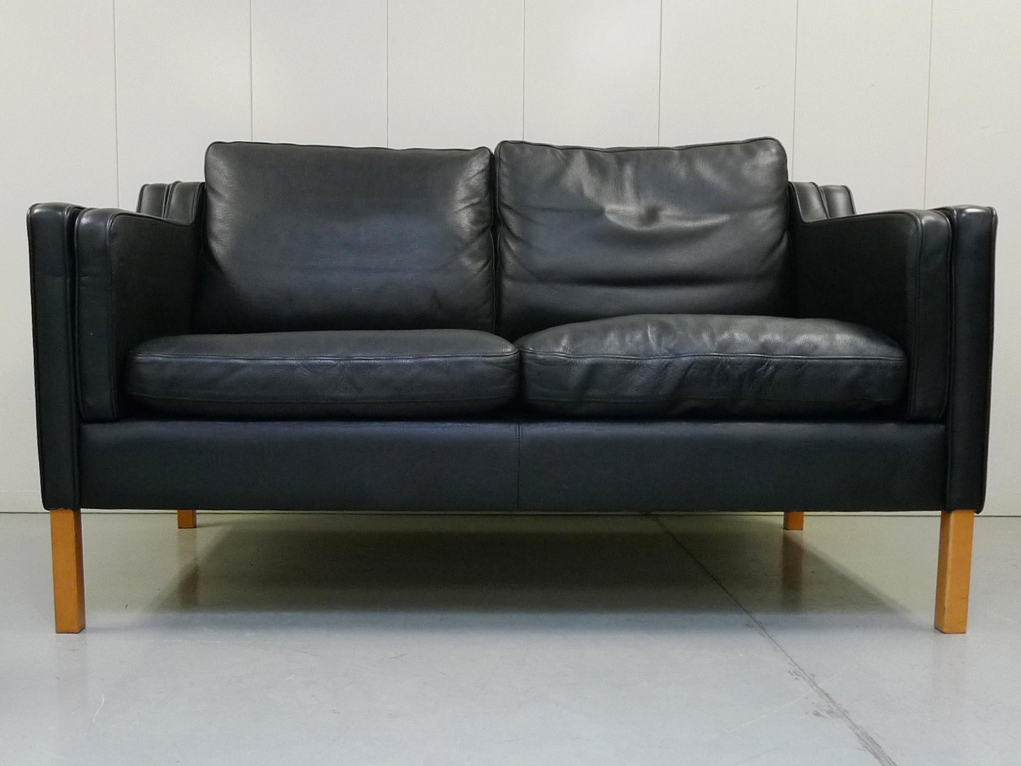 This Minimalist leather settee was designed in the manner of Børge Mogensen and manufactured by Stouby. Its design is one of clean lines and soft, rounded edges. The upholstery is a well-aged black leather, while the frame is beechwood. The settee