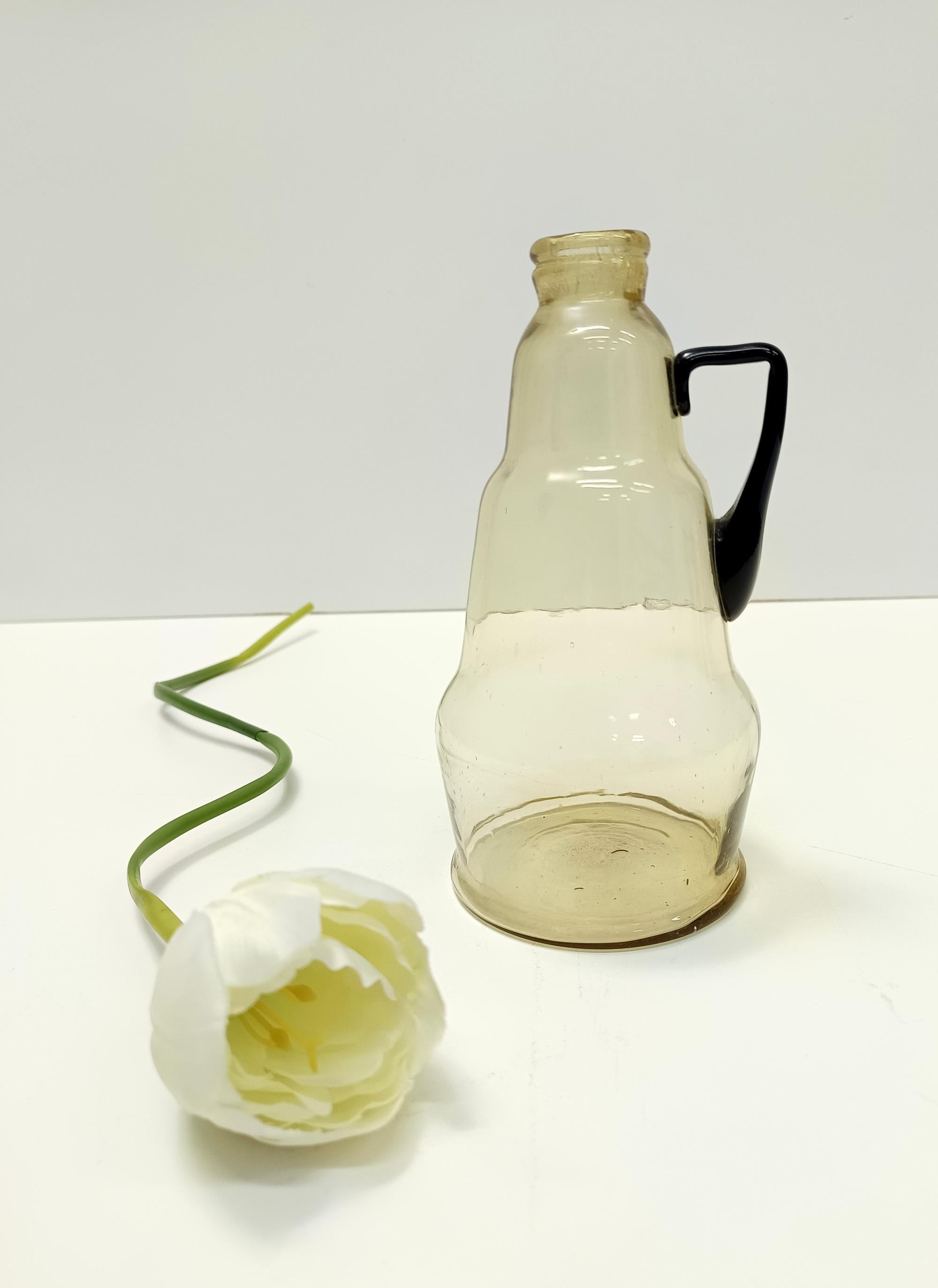 Prodotto in Italia, anni '10. 
Questo vaso a brocca è realizzato in vetro soffiato pagliesco trasparente e la superficie esterna è leggermente rigata. 
Presenta un manico in vetro di Murano nero. 
Si tratta di un articolo vintage, quindi potrebbe