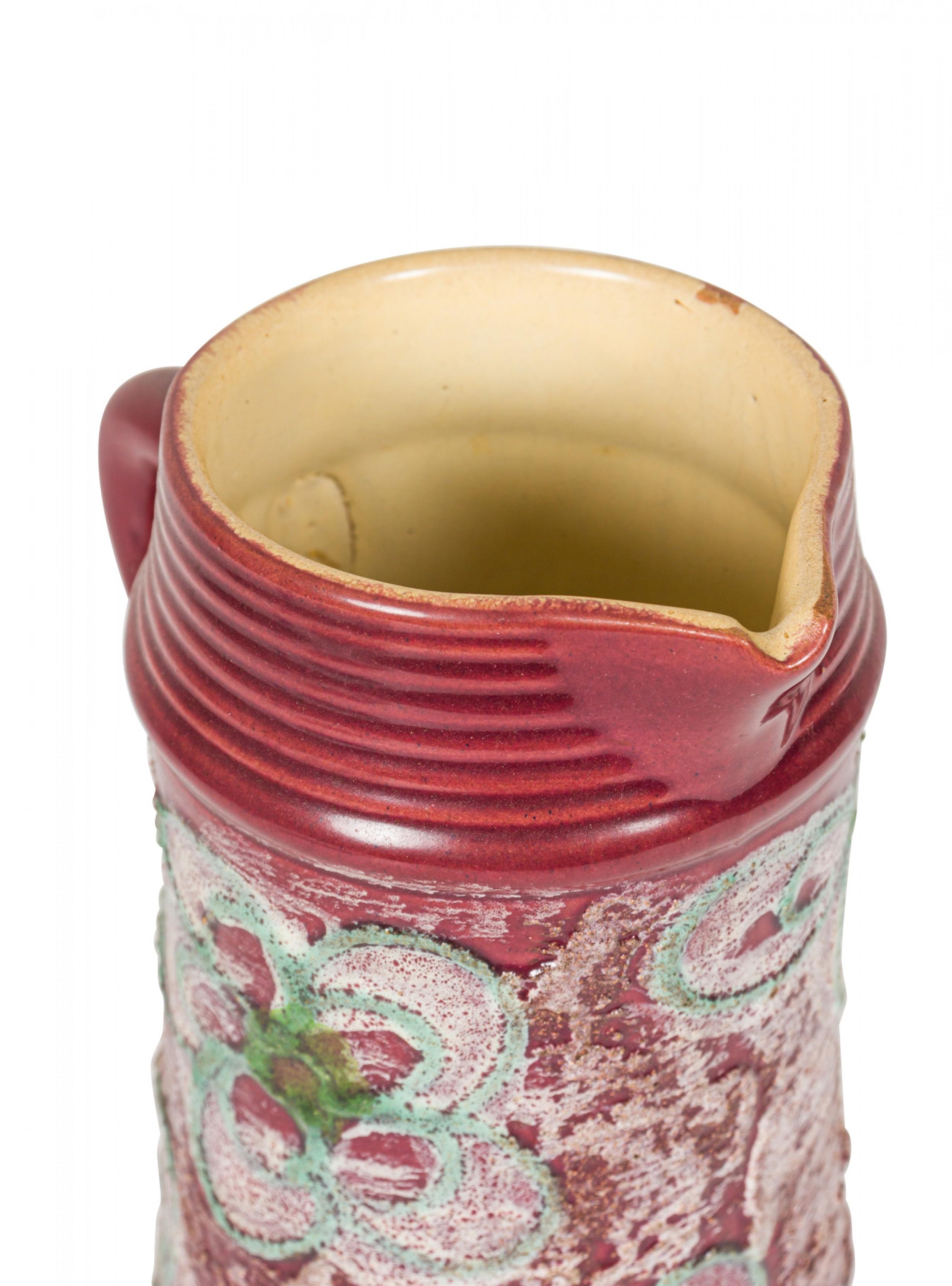 Brocca in ceramica della Germania dell'Est della metà del secolo scorso con un disegno floreale astratto in rilievo di colore beige e verde su un fondo smaltato di colore bordeaux con un motivo a coste intorno alla base e alla bocca della brocca.