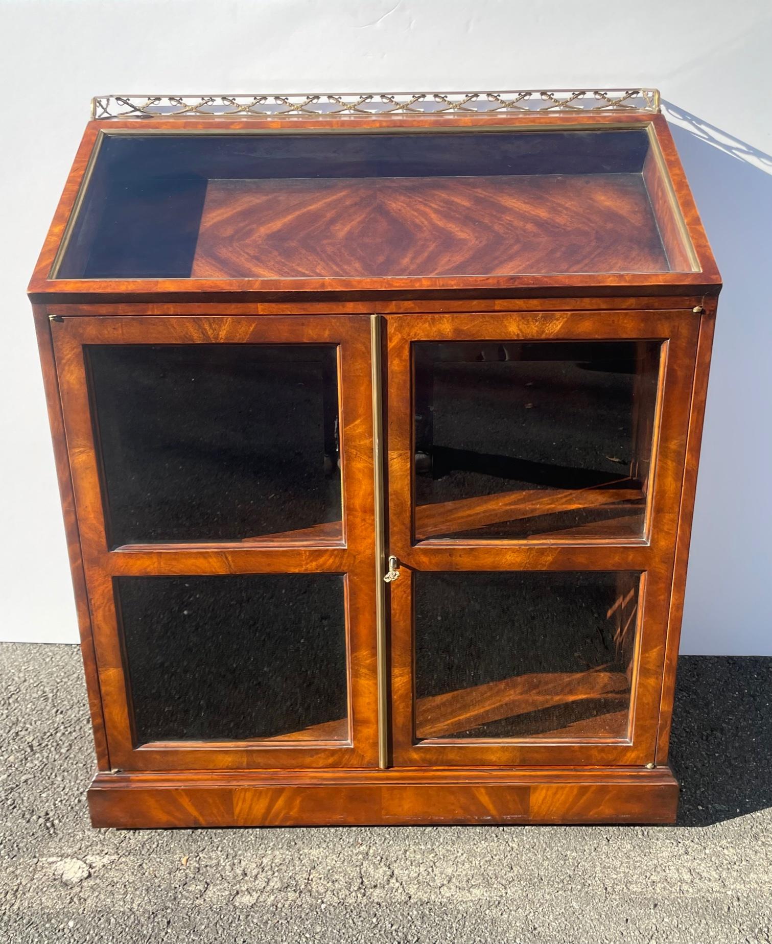 Striking book matched mahogany lecturn cabinet with handsome gold lion hardware and brass gallery with sword motif. There are two glass panel doors that open to reveal tooled leather wrapped shelves. The slanted top is great place to display a