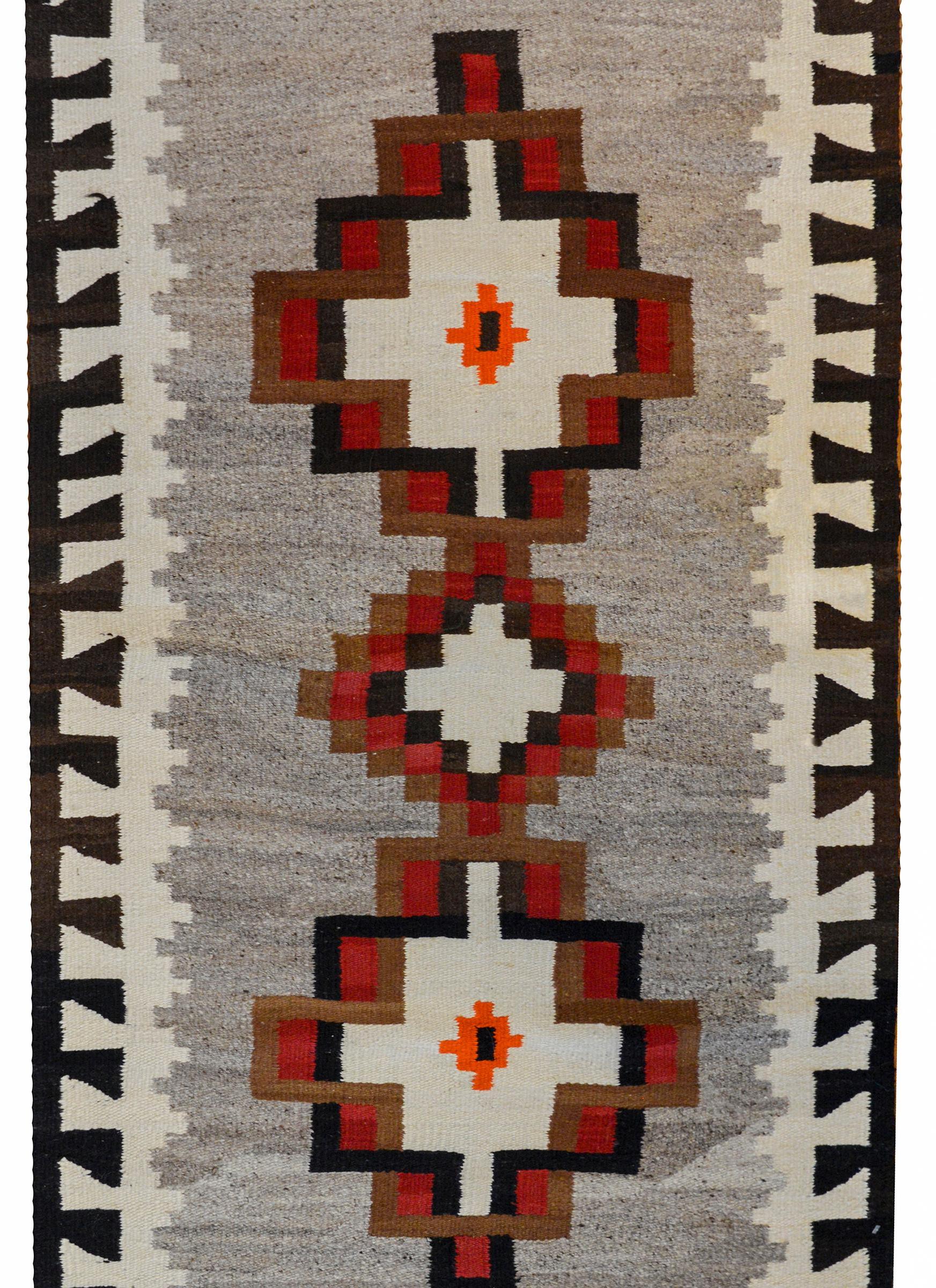 A striking early 20th century Navajo rug with two large diamond medallions flanking a smaller diamond medallion, all woven in crimson, brown, black, and white wool on a gray background. The border is wonderful with a geometric pattern woven in black