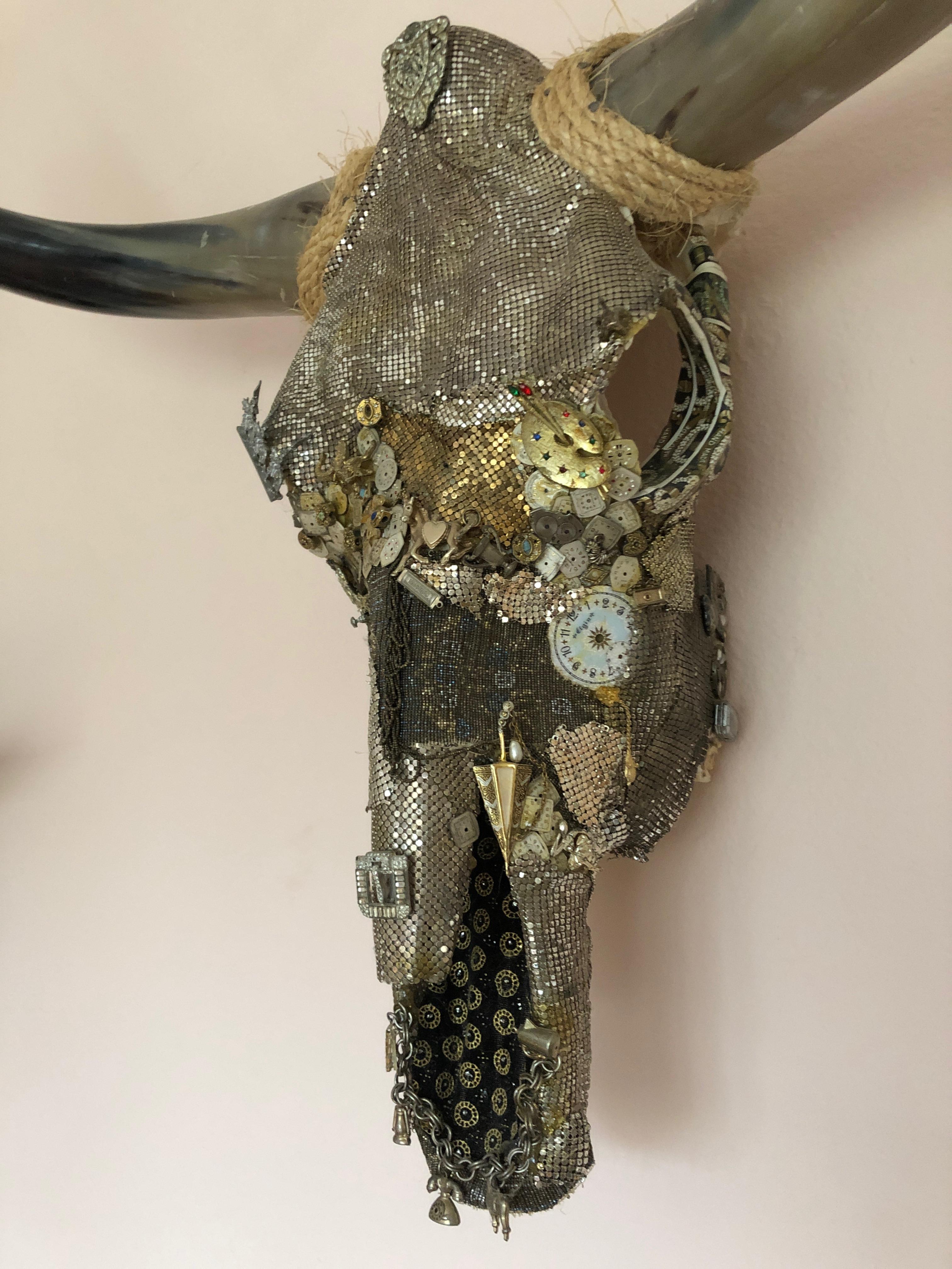Fantastic mixed-media collage on a large longhorn skull that glitters with silver mesh from vintage handbags and is bejewelled with intricate layers of watch faces, charms, a rhinestone shoe buckle, in a masterful mix of silver, black and gold.
By
