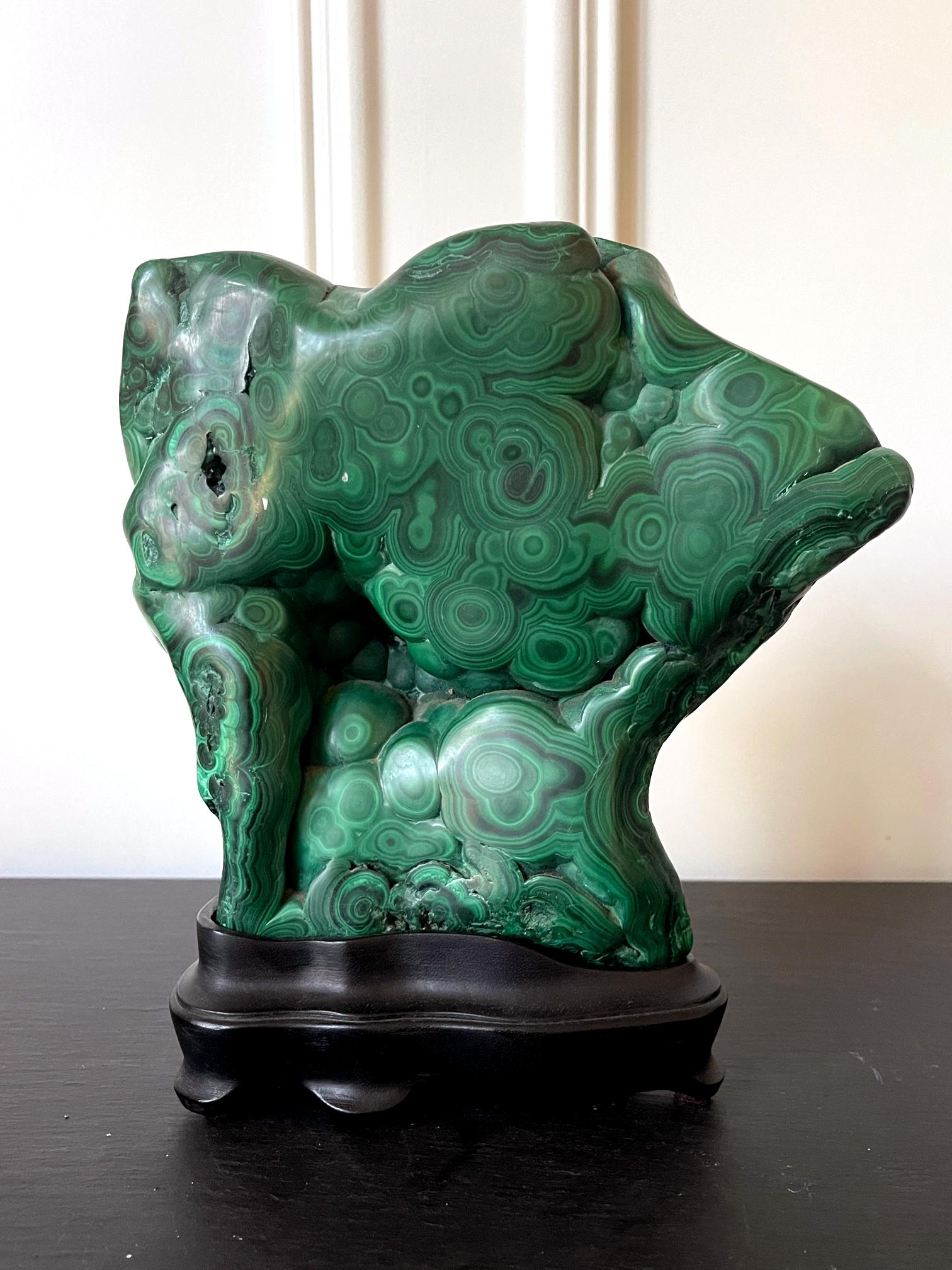 A natural malachite rock specimen in form of an upright slab with striking green and black colors fitted on a wood stand for viewing and meditating. The front and sides of the gemstone feature tight and small polished botryoidal form, revealing a