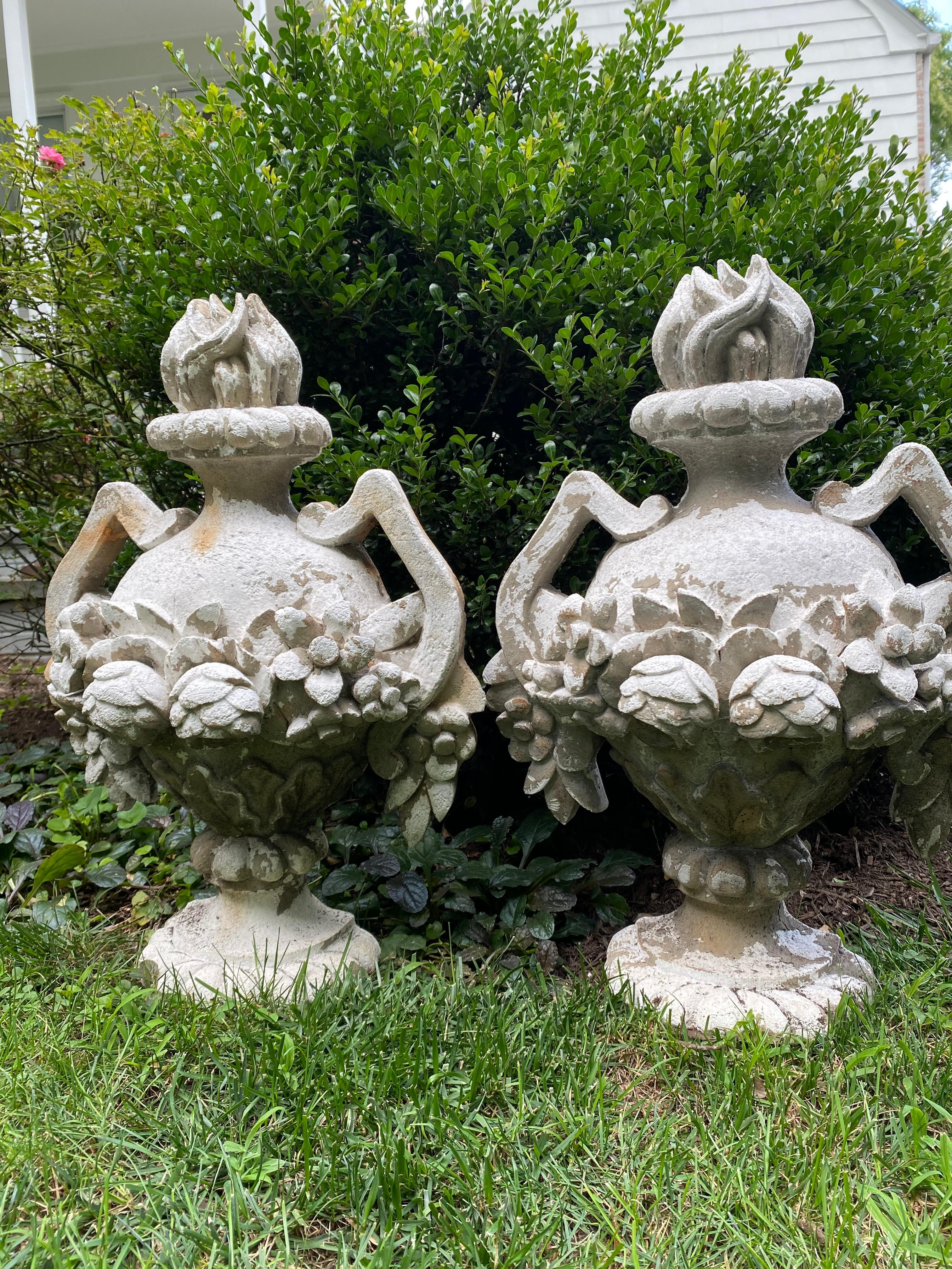 A charming pair of composition and plaster garden decorations in the shape of neoclassical urns having roses on the body, handles and flame shaped finials. The urn shapes are hollow on the reverse sides and flat.  There are metal bars on the backs