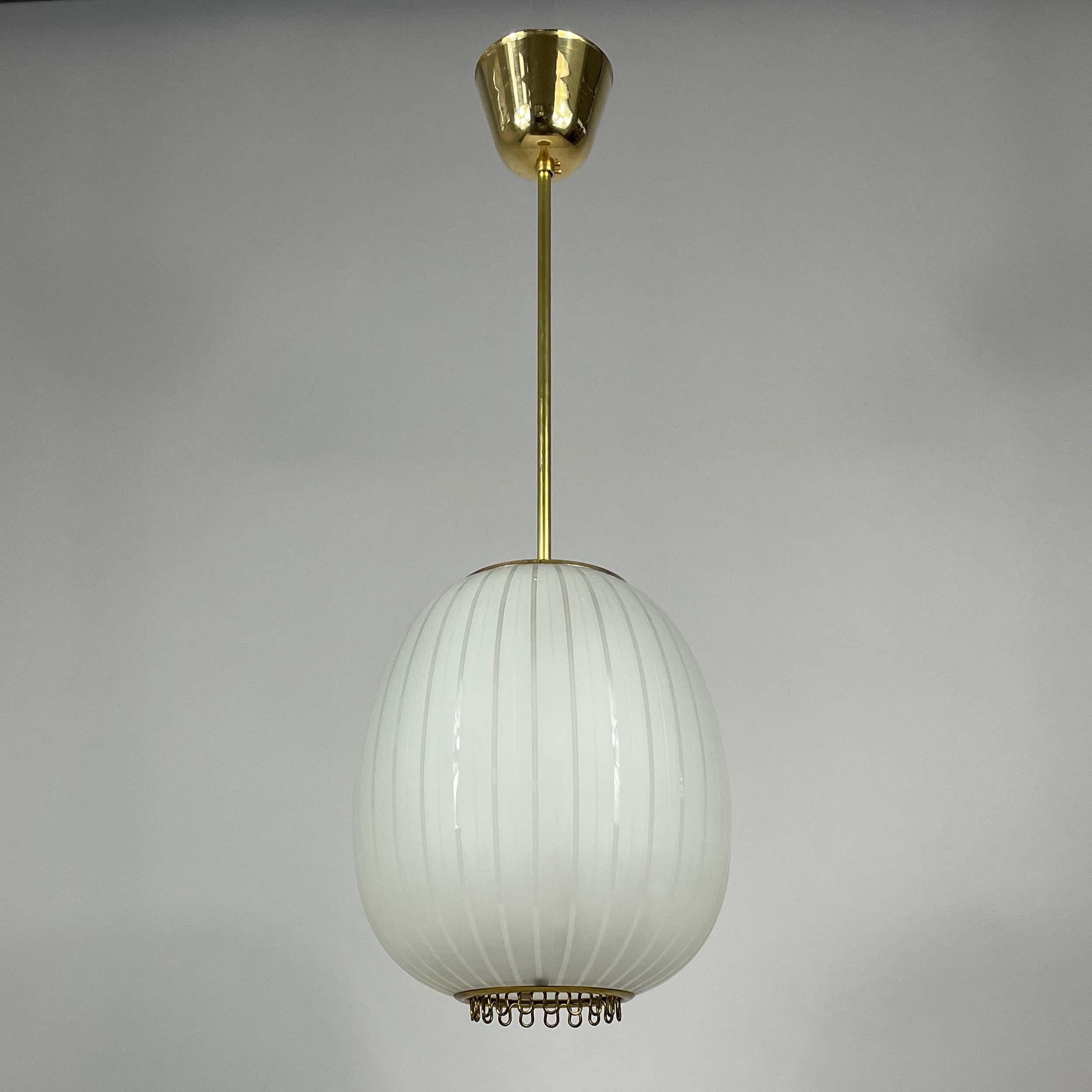 This unusual mid-century pendant was designed and manufactured in Sweden in the 1950s. 

The light features a combination of brass and a white striped frosted glass lampshade. At the bottom there is a brass ring with small decorative loops, adding a