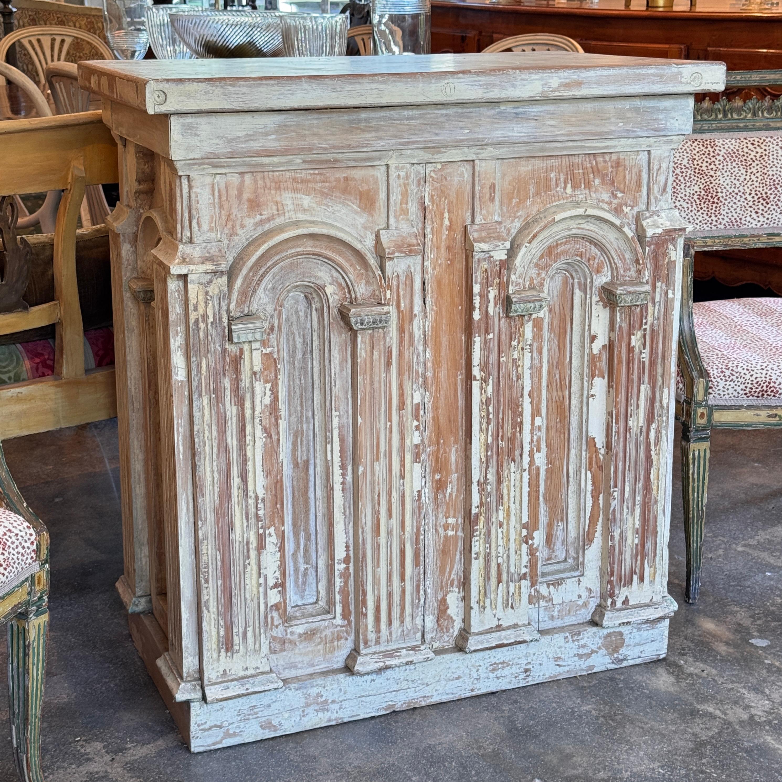Mueble consola con acabado decapado que revela restos de pintura antigua. La fachada presenta paneles arquitectónicos profundamente tallados con motivos arqueados, que le confieren una fuerte presencia arquitectónica. La superficie desgastada