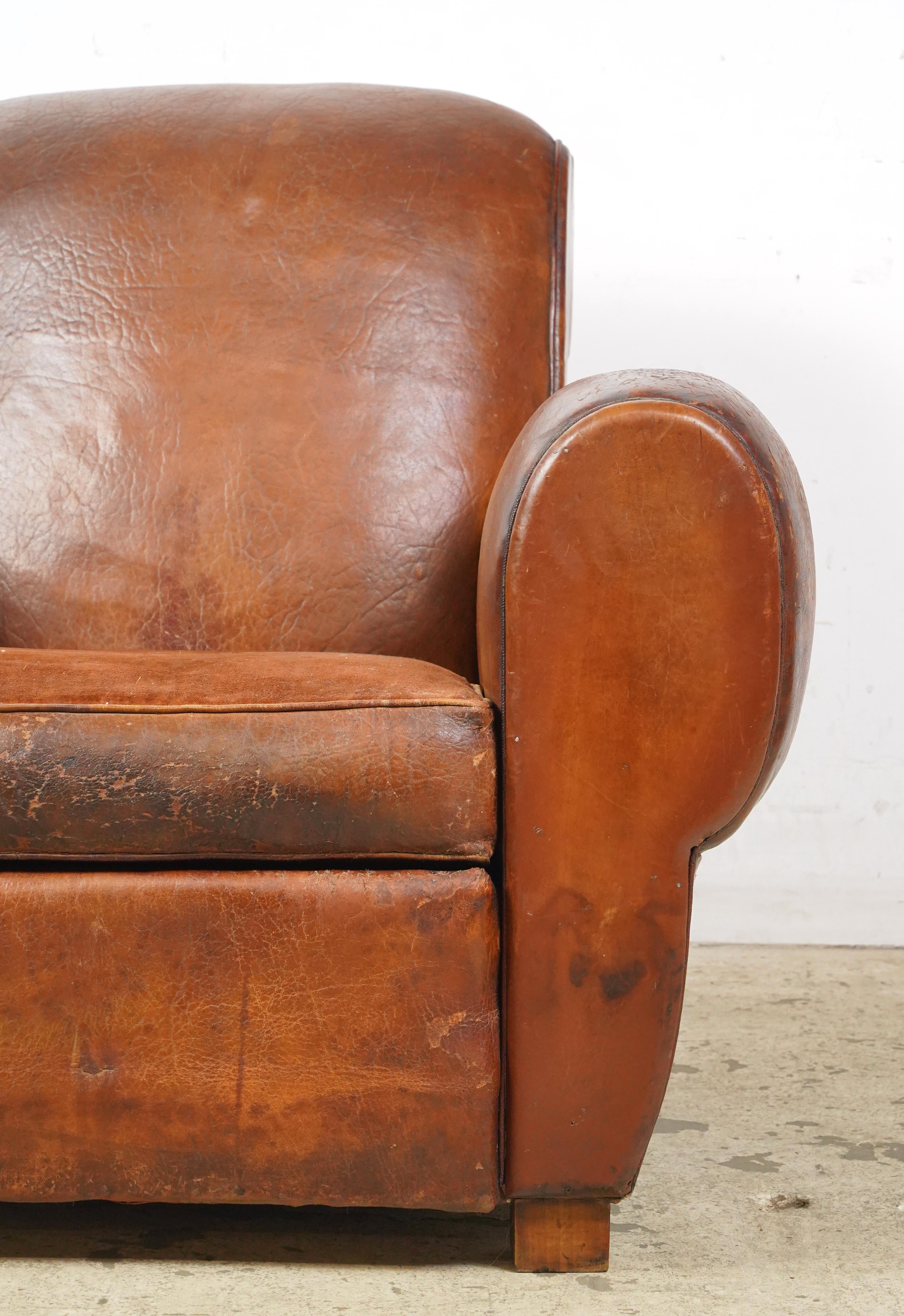 Dark brown leather club chair made in France. It has a low back, wide armrests, a deep cushioned seat, a studded back, and wooden block legs. The leather and feet show some signs of wear and age. Please refer to the pictures for a full view of the