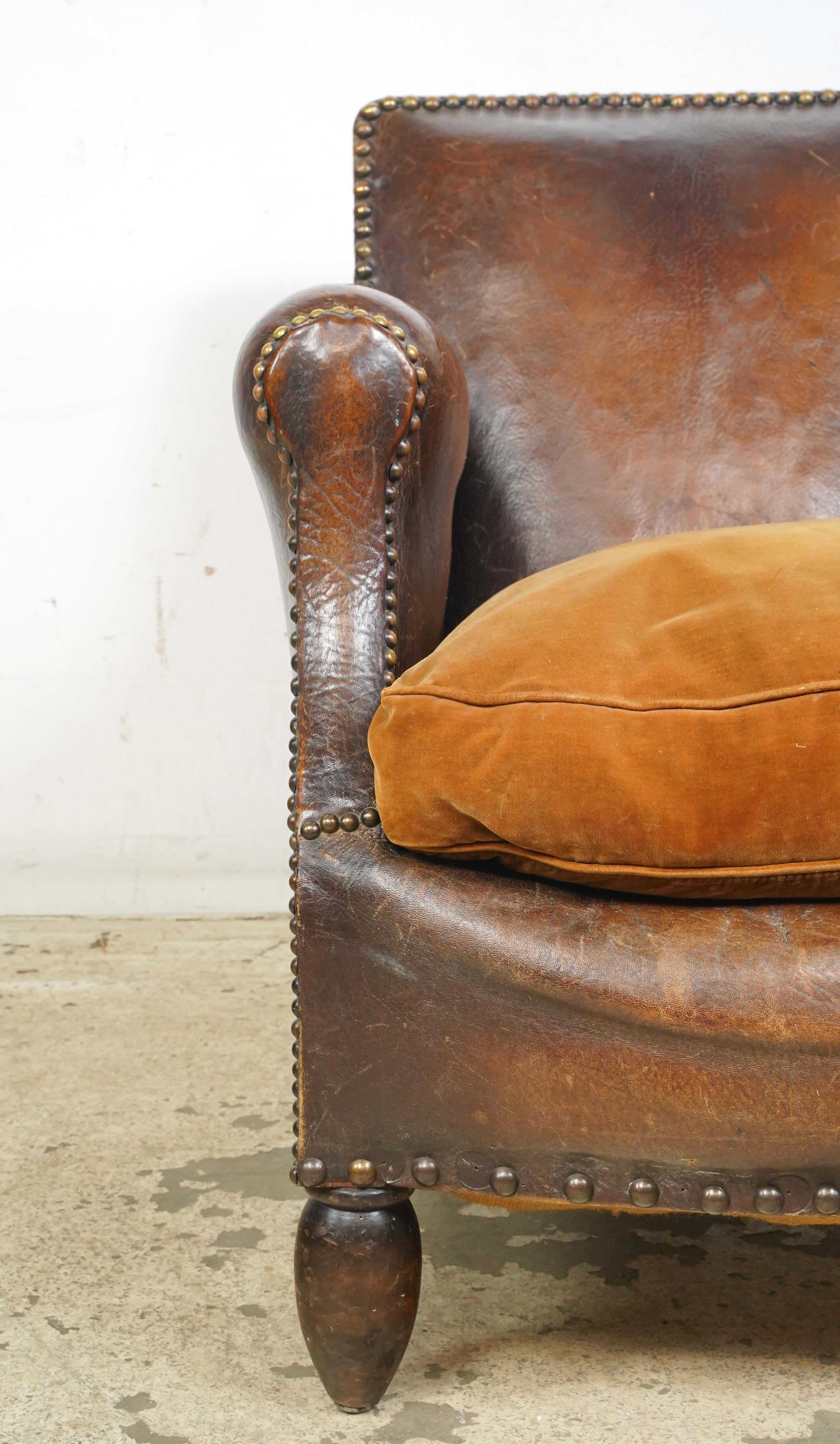 Dark brown leather studded club chair made in France. It has a rounded back, wide armrests, a plush light brown cushioned seat, studded details, and wooden legs. The leather and feet show signs of wear and age. Please refer to the pictures for a