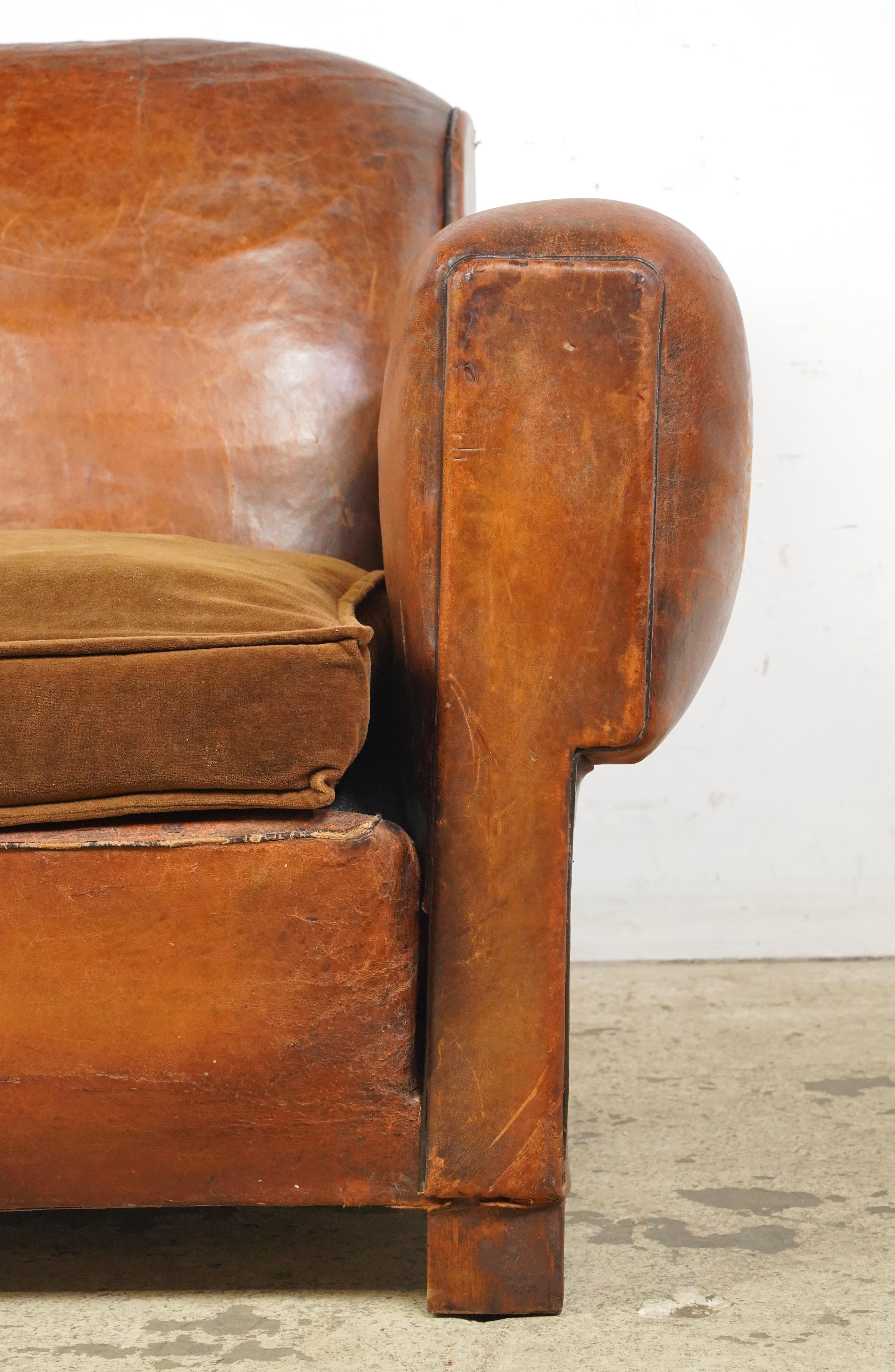 Dark brown leather club chair made in France. It has a low back, wide armrests, a deep cushioned seat, studded details, and wooden block legs. The leather and feet show signs of wear and age. Please refer to the pictures for a full view of the