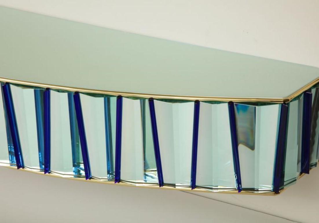 Console murale flottante revêtue de panneaux de cristal aqua-marine clair taillés et polis à la main, alternant avec de fines ailettes en verre bleu. Forme à front arqué, plateau en verre opaque et garniture en laiton. Signature de l'artiste sur le
