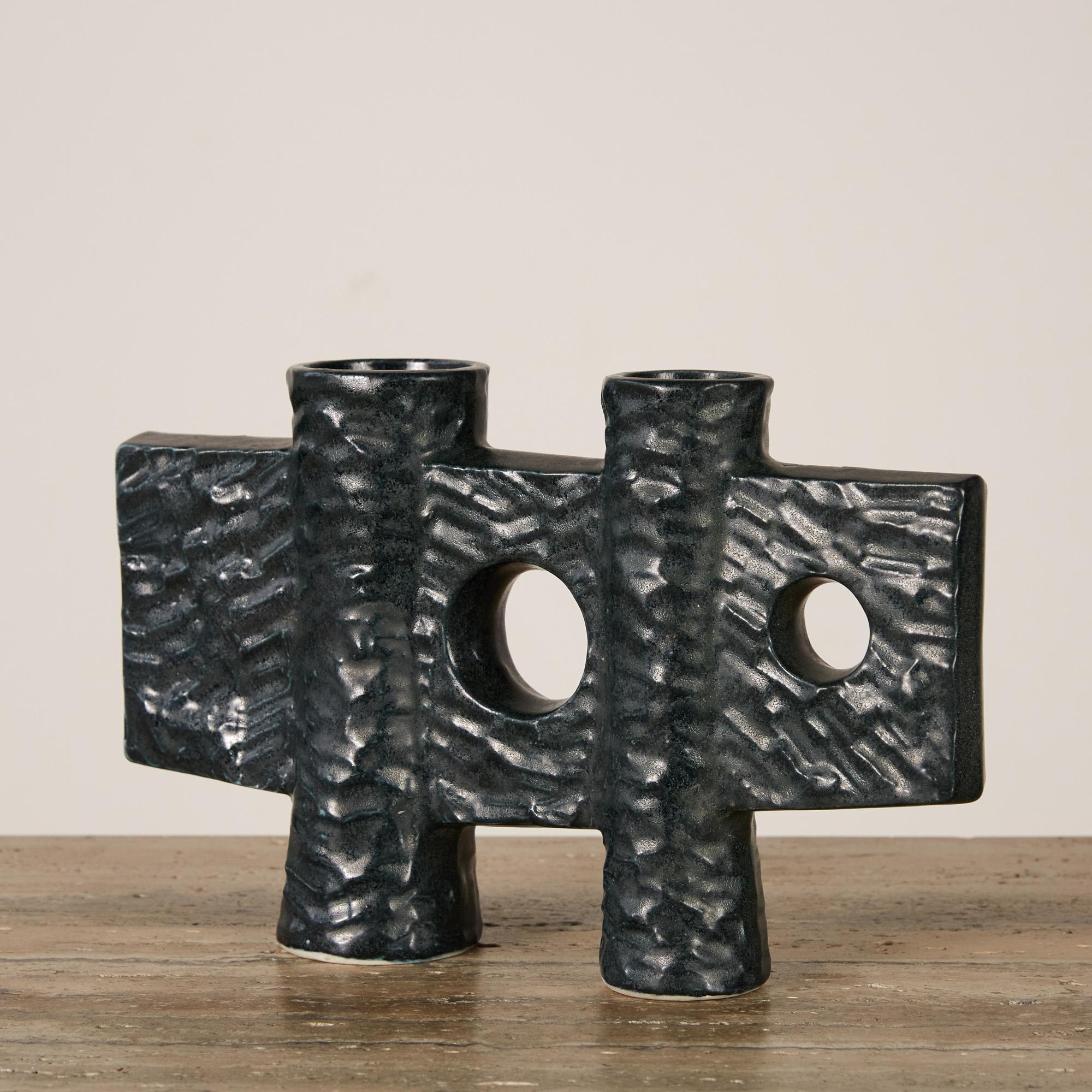 A unique piece of ceramic art that also serves as a vessel. It features a textured greenish-black glossy glaze with two cylindrical structures that are connected by a flat vertical rectangular form with round cutouts in the center Perfect for