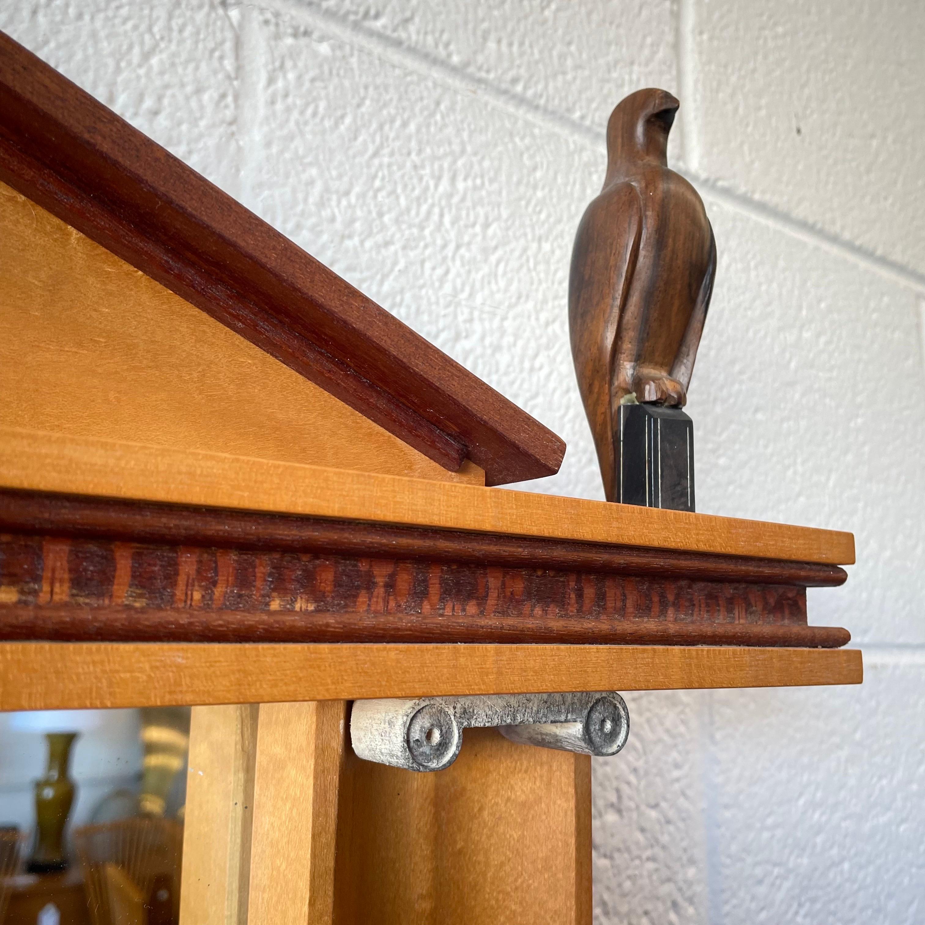 Miroir Vintage Studio craft en divers bois exotiques. Le miroir est conçu avec  colonnes en ruine et un aigle en bois de rose sculpté à côté du fronton.

Signé par l'artiste au verso mais illisible.