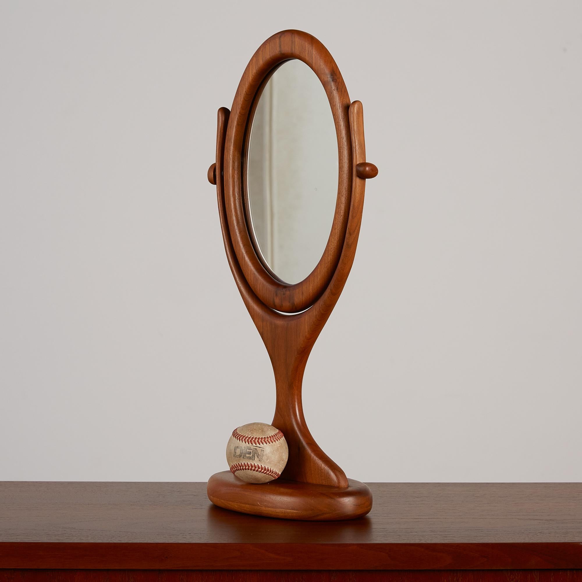 This hand carved vanity mirror features a thick sculpted walnut frame with an oval shaped piece of inset glass. The mirror is set in a swivel frame to adjust your angle. This playful design would be perfect for a vanity, or on a shelf or tablespace