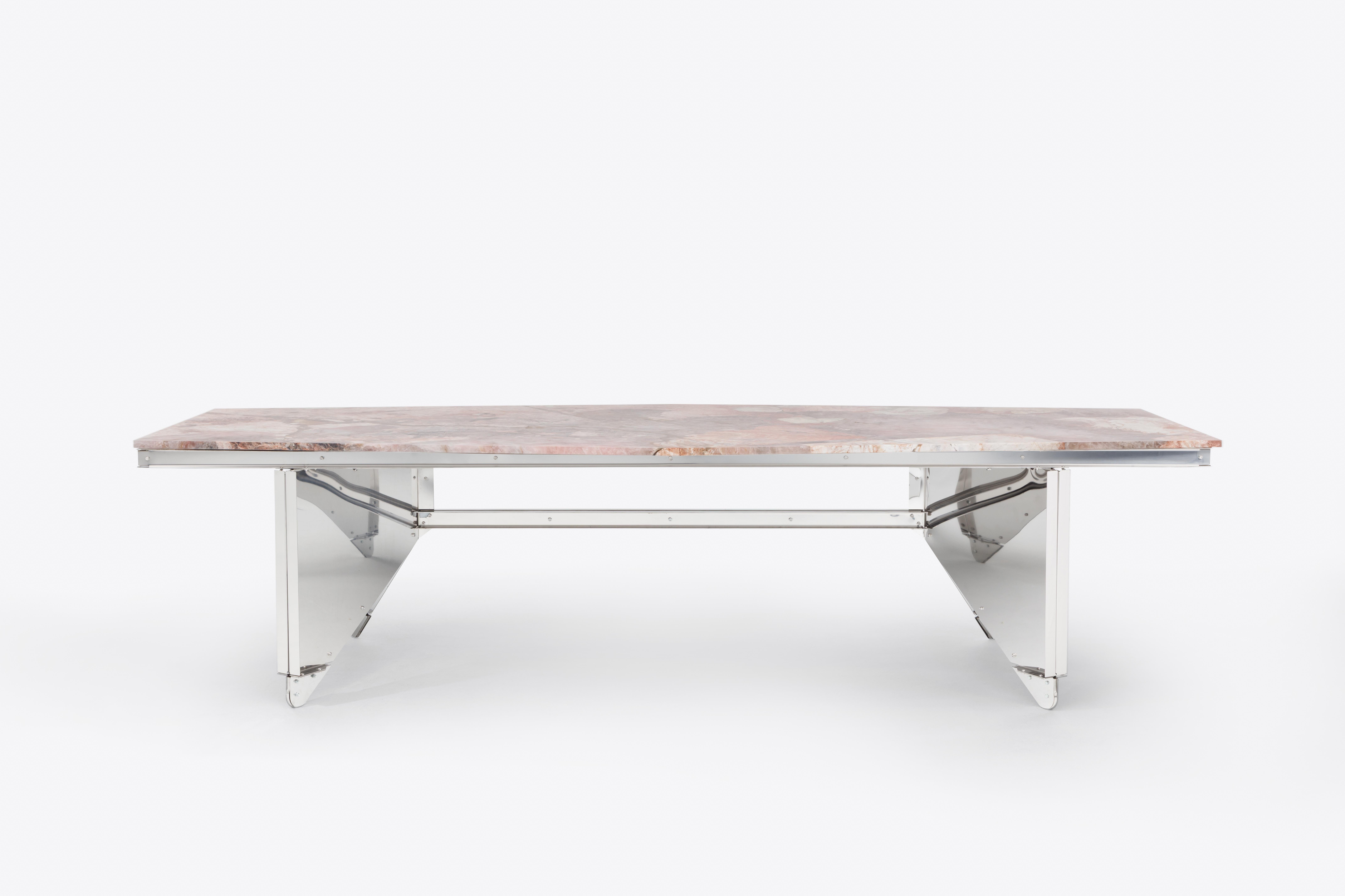 This dining table contrasts a precisely engineered stainless steel base with a richly veined pink Patagonia quartzite top. The angular structure lifts the heavy stone with unexpected lightness, merging mineral complexity with structural logic—a